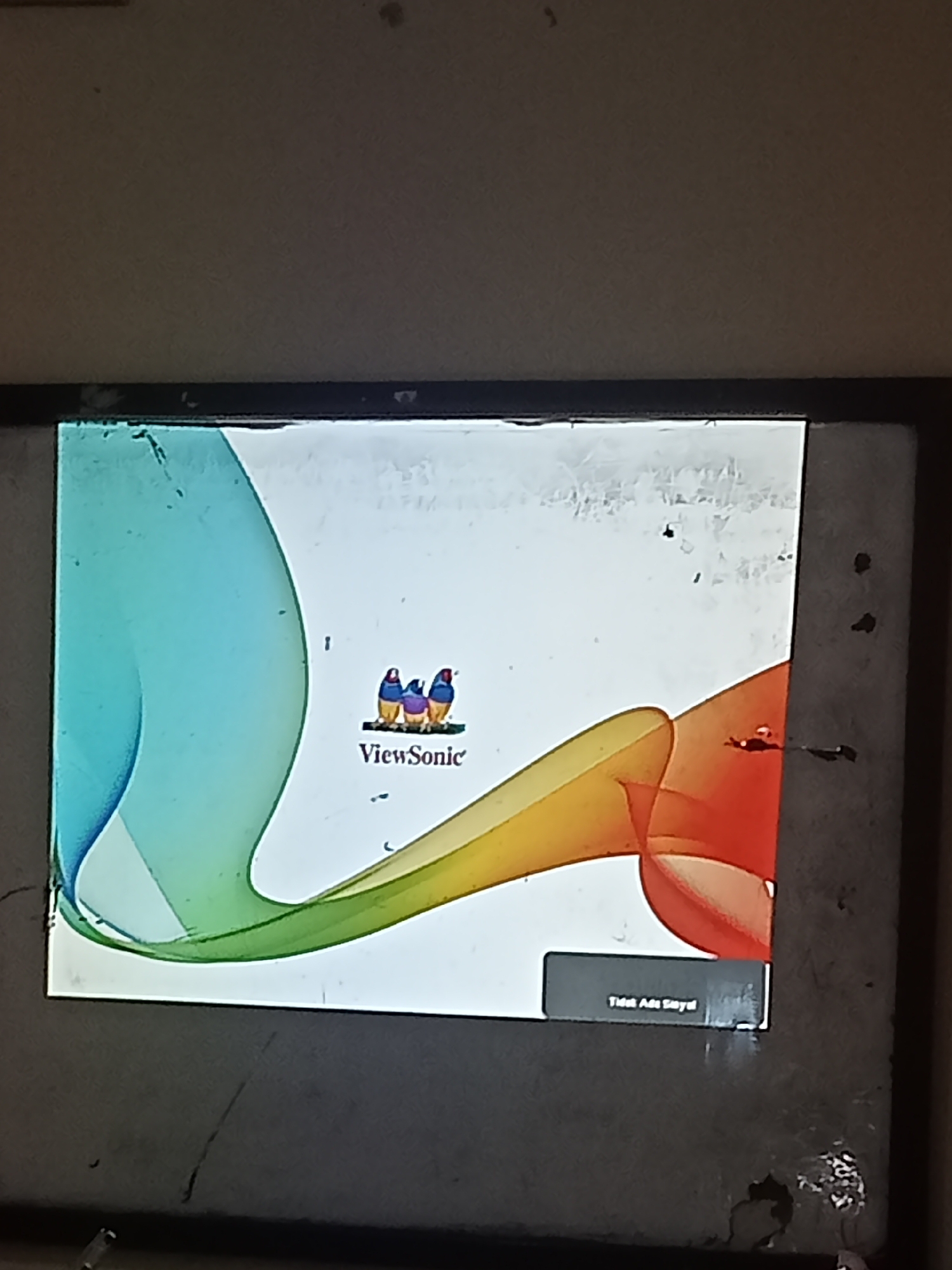 The image shows a projection of the ViewSonic logo and a colorful abstract design on a screen. The logo features three small birds, two blue and one purple, perched on a yellow branch, with "ViewSonic" written below. The abstract design consists of flowing shapes in shades of blue, green, yellow, and orange. A dark rectangular bar with white text, which appears to read "Tidak Ada Sinyal" (which translates to "No Signal" in Indonesian), is visible at the bottom of the screen. The screen itself is framed by a dark, worn bezel. The background behind the screen is a plain, neutral-colored wall with some discoloration and marks, suggesting an indoor environment. There is no indication of the city of Secang, Indonesia, within the image itself.