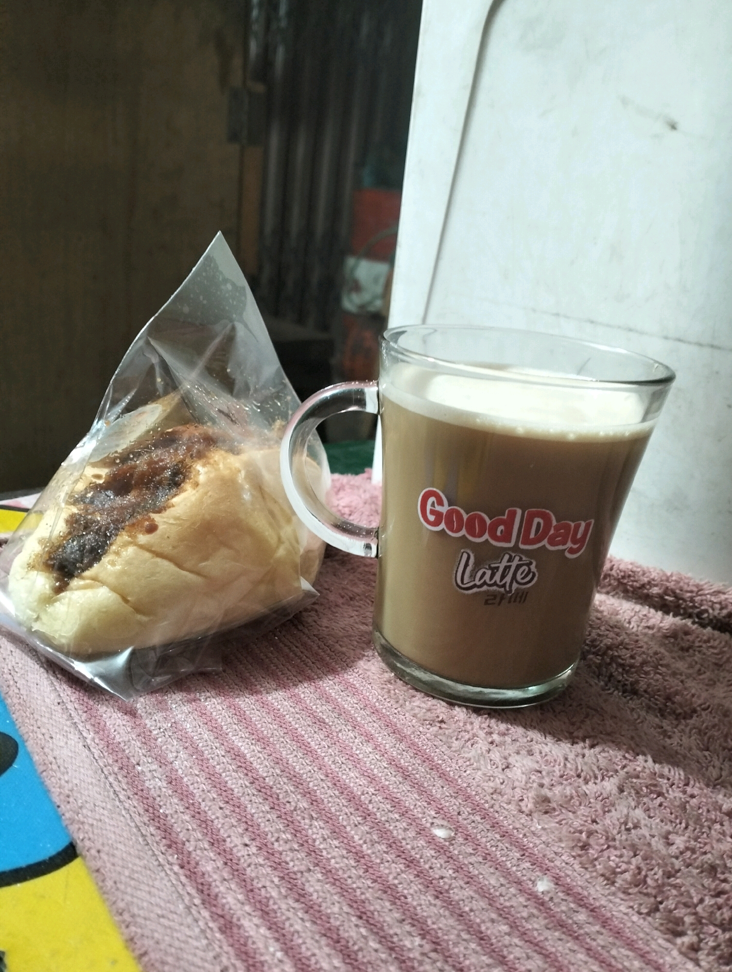 In Tarai Bangun, Indonesia, a close-up shot captures a glass mug with a handle filled with a light brown beverage, likely coffee with milk, topped with foam. The mug has a red and white sticker that reads "Good Day Latte" and a smaller text below it. Beside the mug, a round pastry is partially visible through clear plastic packaging. The pastry appears to have a dark, possibly caramelized or burnt sugar topping. Both the mug and the pastry are placed on a pink and white striped cloth. In the background, the scene is dimly lit, with vertical corrugated metal or siding and a rough, textured wall. The overall setting suggests an indoor, possibly domestic or casual environment.