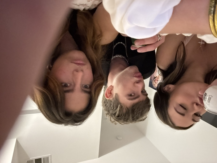 Three individuals pose close to the camera, which is tilted upside down. A woman with long brown hair and a stern expression is to the left. In the center, a male with light hair makes a kissing face. A woman with dark hair, long lashes, and a drink in her hand is on the right. They appear to be indoors, with glimpses of a white ceiling, a vent, and a picture on the wall visible. The lighting is dim, giving the image a somewhat intimate feel.