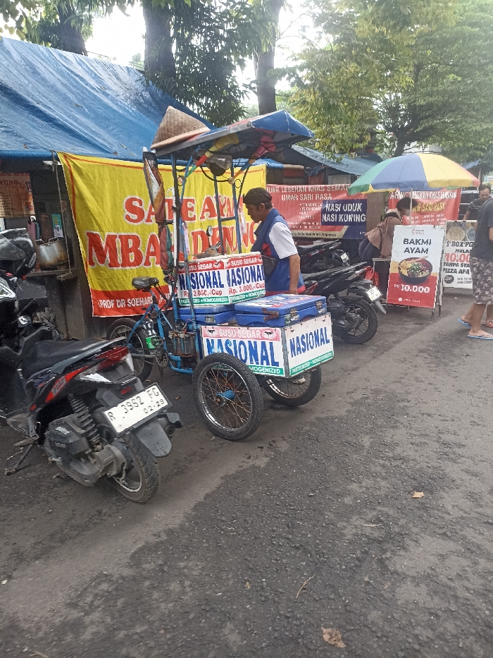 The scene takes place on a street in Purwokerto, Indonesia, located at coordinates -7.417 latitude and 109.249 longitude. A man in a white and blue vest is standing next to a food cart displaying a sign with "SUSU SEGAR NASIONAL" written on it. The cart is situated in front of a building with a blue tarpaulin roof. Two motorcycles are parked on the left side of the frame. A bright yellow sign is partially visible, featuring the text "SARE AYAM MBA DENG", partially obscured by the food cart. To the right of the cart, a person stands near another food cart, this one with a sign displaying the text "BAKMl AYAM 10.000". The road is paved with gray asphalt and is relatively empty. The sidewalk has trees and other buildings visible in the background. The scene appears to be in front of a restaurant or food stall, perhaps a market. It's likely daytime and the weather is clear.