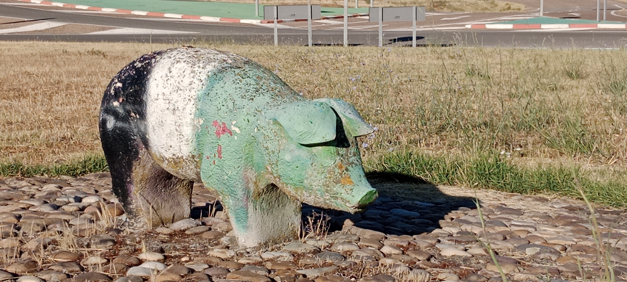 An outdoor, medium shot captures a weathered, statue of a pig in Oropesa, Spain. The statue is predominantly painted a faded green with patches of white and black on its rear. It rests on a bed of small, rough stones, with dry, straw-like grass surrounding it. In the background, a paved area with red and white curbs suggests a road or track, possibly a race circuit. Several metal poles with grey rectangular panels are also visible in the background, along with a hazy, sunlit sky. The scene is set during the daytime, under bright, natural light, casting a shadow of the pig statue onto the stony ground. There are no people or animals present in the image.