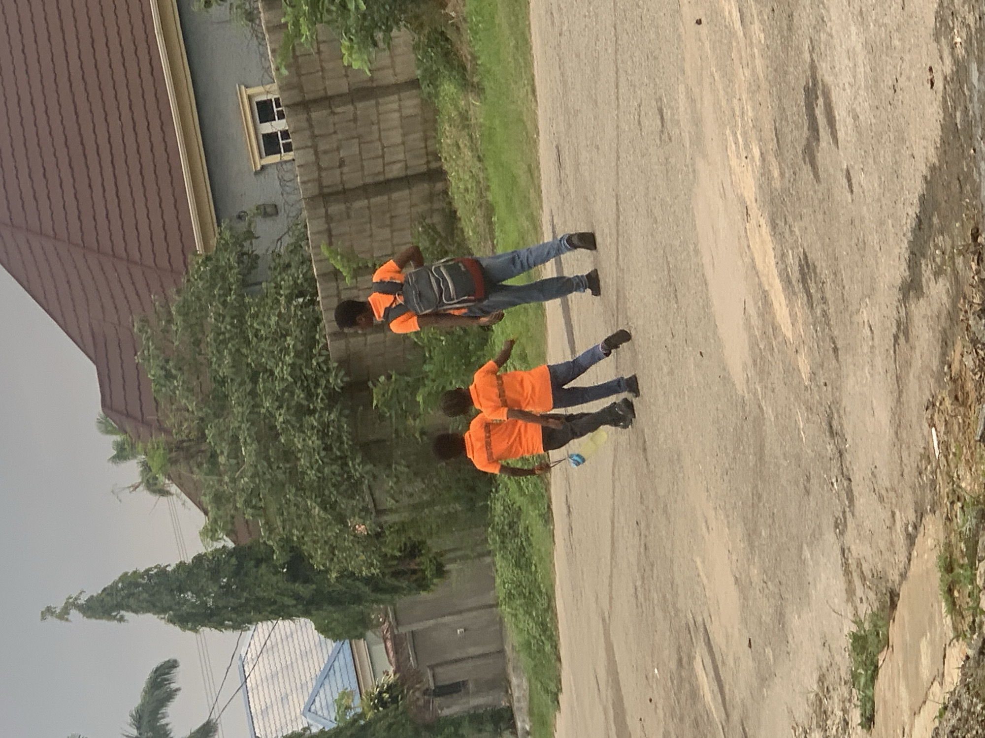 In an outdoor, paved setting that appears to be a residential street or road in Abuja, Nigeria, three individuals, two boys and a young man, are depicted walking away from the camera. The young man, positioned slightly ahead and to the right, wears an orange t-shirt and blue jeans, carrying a dark backpack with red accents. The two boys are dressed in matching orange t-shirts and dark pants. The boy on the left is holding a blue object and a yellow plastic bottle. The background reveals a gray, brick-like wall topped with barbed wire, suggesting a secure perimeter, and beyond that, houses with brown and blue roofs, interspersed with lush green trees and vegetation. The sky is overcast, indicating a cloudy or possibly rainy day. The ground is paved with asphalt, showing signs of wear and tear with visible patches and some scattered debris. The overall scene suggests a casual outing or perhaps a walk home.