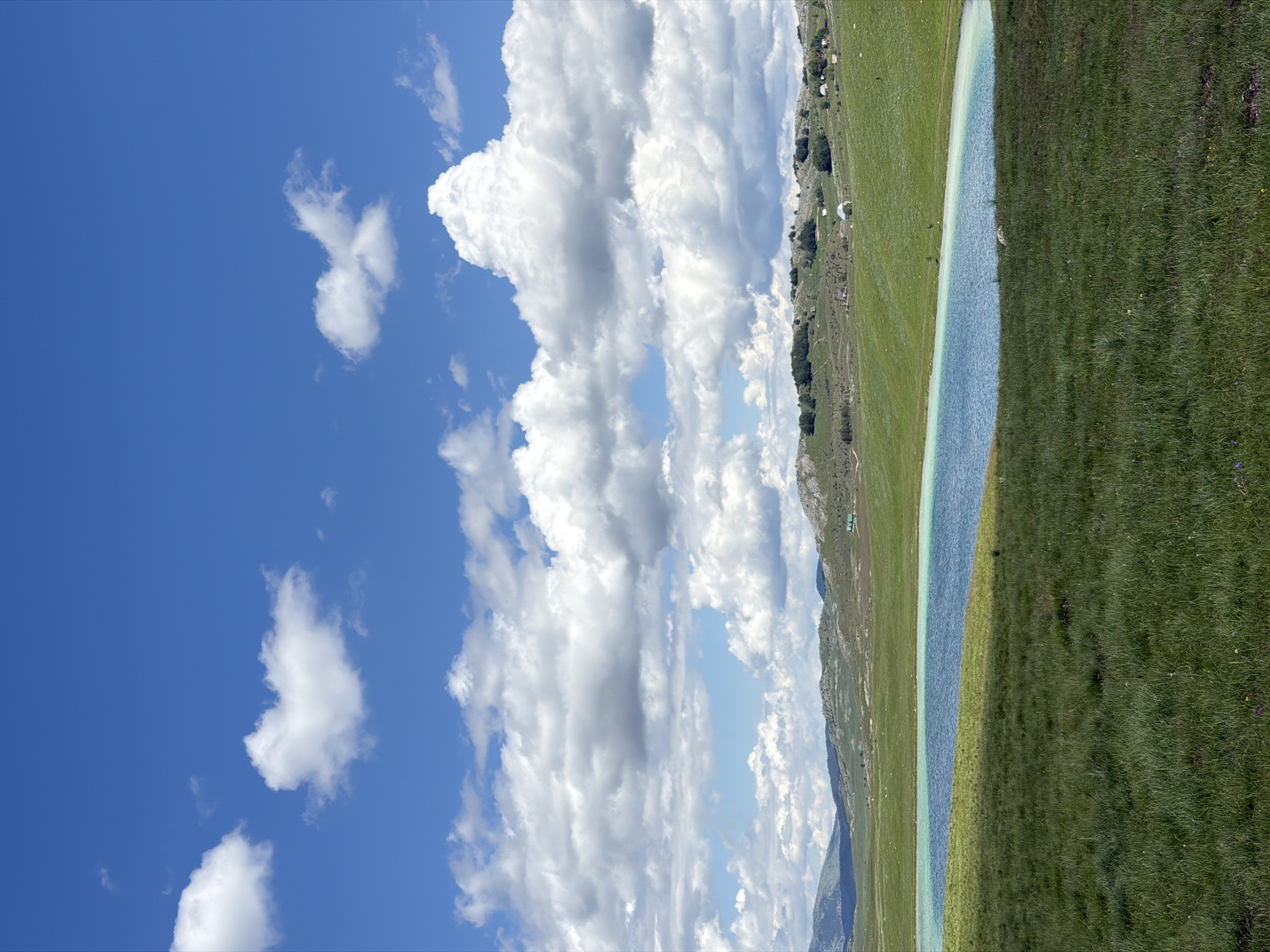 This detailed description pertains to an outdoor landscape photograph taken in Novakovići, Montenegro. The image showcases a natural environment under a bright, partly cloudy sky. The upper portion of the frame, approximately 60%, is occupied by a clear blue sky dotted with numerous white cumulus clouds of various sizes and densities. Below the sky, a series of gently rolling hills, covered in lush green vegetation, extends across the mid-ground. Scattered across these distant hills, particularly on the right side, are small, light-colored structures, likely houses or agricultural buildings. In the mid-ground, an irregularly shaped body of water with vibrant blue and turquoise hues is visible, its surface calm and reflecting no distinct objects. The water exhibits a lighter, almost aqua color along its nearer edge, transitioning to a deeper blue. The foreground, occupying the lower approximately 30% of the image, consists of a vast expanse of dense, verdant green grass, suggesting a well-maintained meadow or pasture. There are no individuals, animals, or vehicles explicitly visible within the frame. The overall impression is one of a serene, rural landscape under daylight conditions.