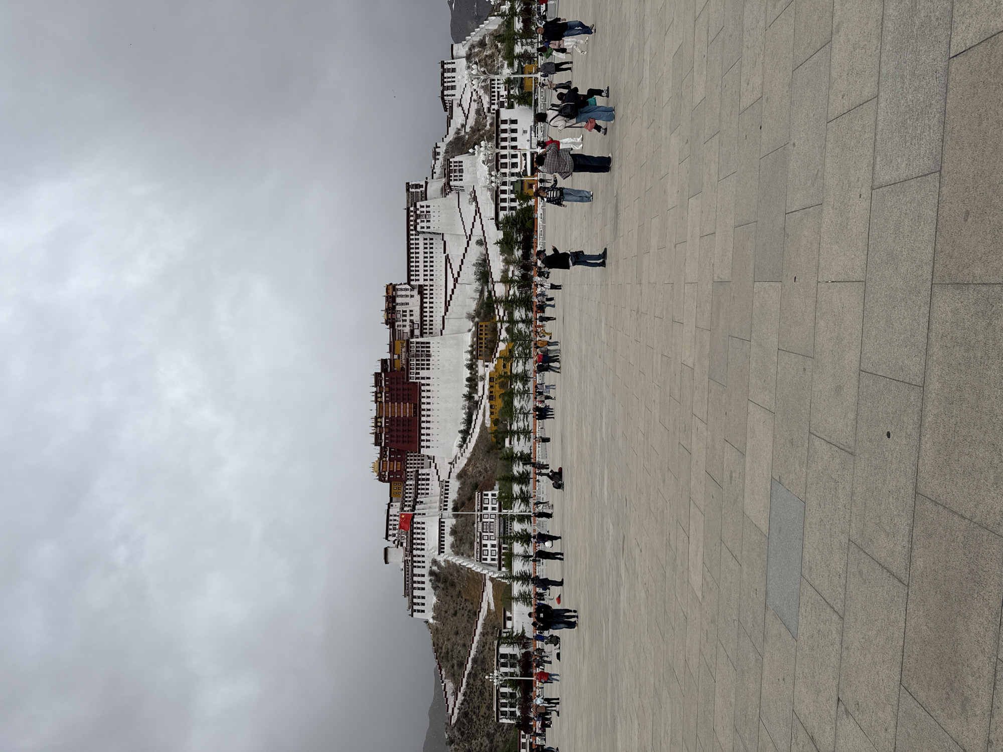 The image shows the Potala Palace in Lhasa, located in the Chengguan District of China. The large, tiered white and red building complex is built on a hillside and stands against a cloudy, gray sky. The foreground shows a large, paved plaza filled with people, some standing and some walking. The people are dressed in a variety of clothing. A row of trees and lampposts lines the edge of the plaza adjacent to the palace. A Chinese flag is visible flying from one of the lower structures of the Potala Palace.