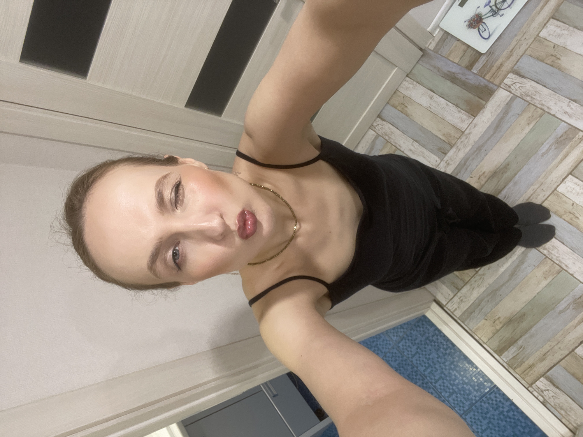 A young woman is taking a selfie in what appears to be a bathroom. She is wearing a black sleeveless dress and a delicate gold necklace. The background features a light-colored door with dark stripes, wooden herringbone patterned flooring, and a glimpse of blue tile flooring in the corner, suggesting the presence of a shower or bath area. The lighting seems to be artificial, coming from overhead. The woman is looking at the camera with a slight pout. There is a chopping board on the floor. It is not possible to confirm if the location is Chaikovsky, Russia based on the image alone.