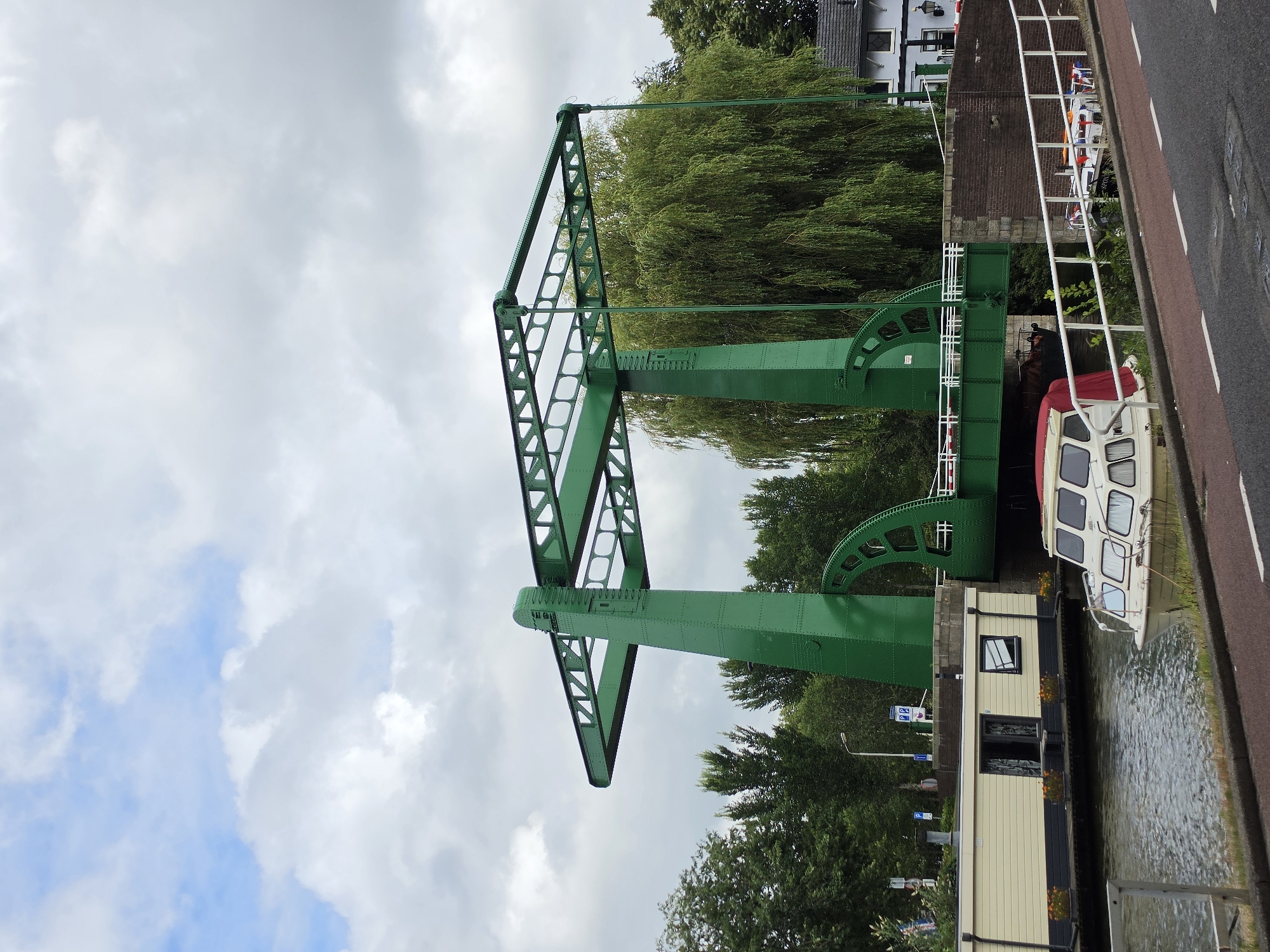 A green metal bridge, likely a bascule bridge, is the central feature of this image, spanning a canal in Delft, Netherlands. The bridge is painted a vibrant green and has a complex structural design with visible beams and rivets. It is in the closed position, allowing passage underneath. On the canal, a long, light-colored houseboat is moored to the left, its windows and door visible. In front of the houseboat, a smaller white boat with a red cover is docked. The water in the canal is dark and calm.
To the right of the bridge, a brick embankment leads up to the bridge deck. Beyond the embankment, a weeping willow tree with dense, green foliage dominates the background. To the right of the tree, a white building with a dark roof and windows is partially visible. The scene is set under a cloudy sky with patches of blue showing through. The ground in the foreground consists of a paved road with white lane markings. The overall impression is of a tranquil canal scene in a European city, with the industrial design of the bridge contrasting with the natural elements and residential structures.