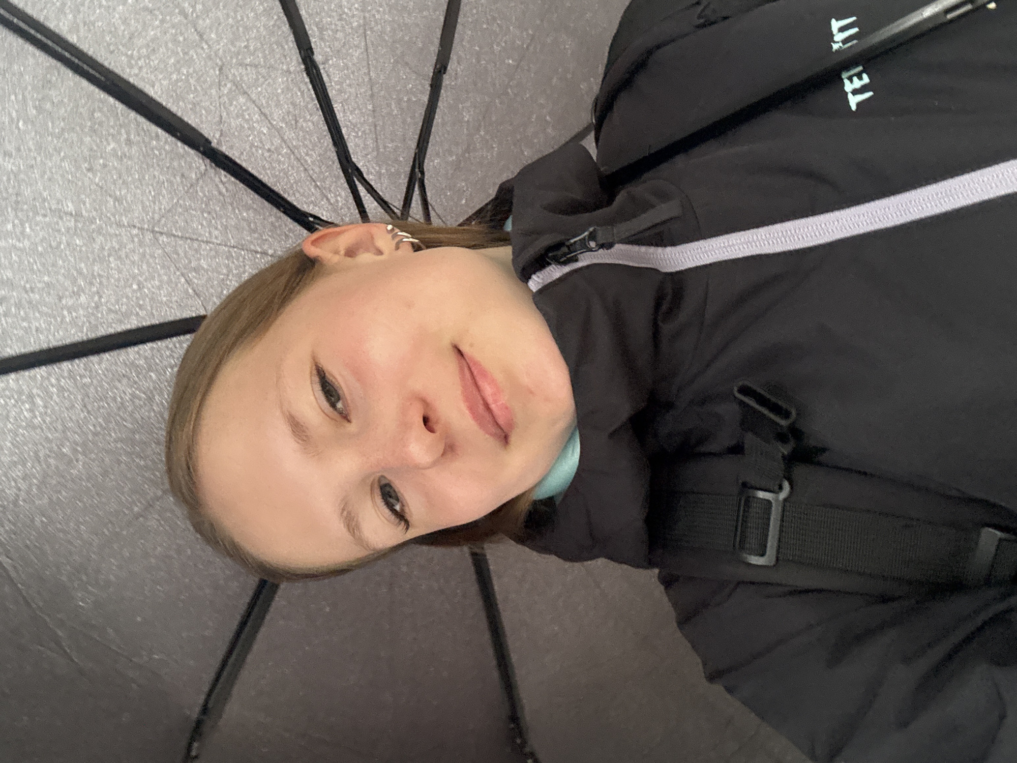 A woman is positioned under an open umbrella, looking directly at the camera. The woman has fair skin, light brown hair, and an earring on her left ear. She's wearing a black jacket with a white stripe near the zipper. A portion of a teal shirt is visible beneath the jacket. The umbrella is dark gray with black support struts. The photo was taken outdoors in Chaikovsky, Russia.
