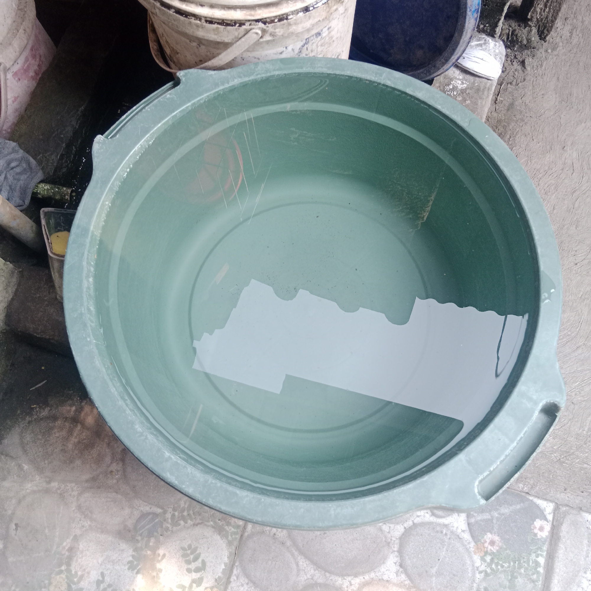 In the city of Tegowanu Kulon, Indonesia, a green plastic tub filled with water sits on a tiled floor. The tub is round with a wide rim and two handles, one of which is visible on the right side. The water inside reflects the overhead light, creating a bright, irregular shape on the surface. Scratches are visible on the inside of the tub. Behind the tub, other containers are partially visible, including a large white bucket with a metal handle and a dark blue bucket. The ground around the tub is made of light-colored tiles with a pattern of stones and some faint floral motifs. To the left of the tub, a dark, wet surface with some debris is visible.