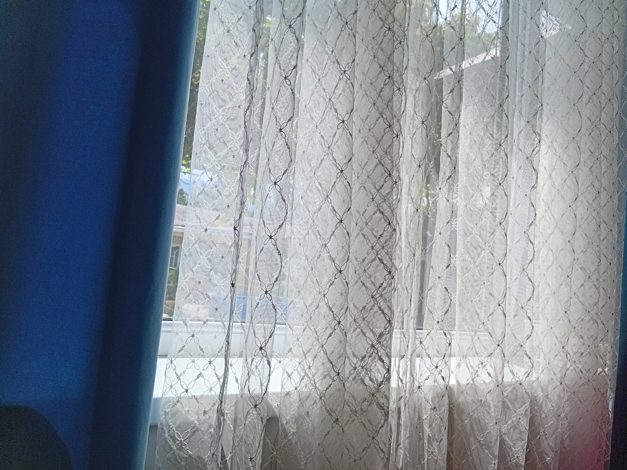 The image shows a window with white lace curtains. To the left of the lace curtains, a dark blue curtain is visible. The lace curtains are patterned with a diamond shape and small floral designs. Through the lace curtains, a window frame and some greenery outside can be seen. The scene appears to be indoors, likely a room with a window. The location is Kadamjay, Kyrgyzstan.
