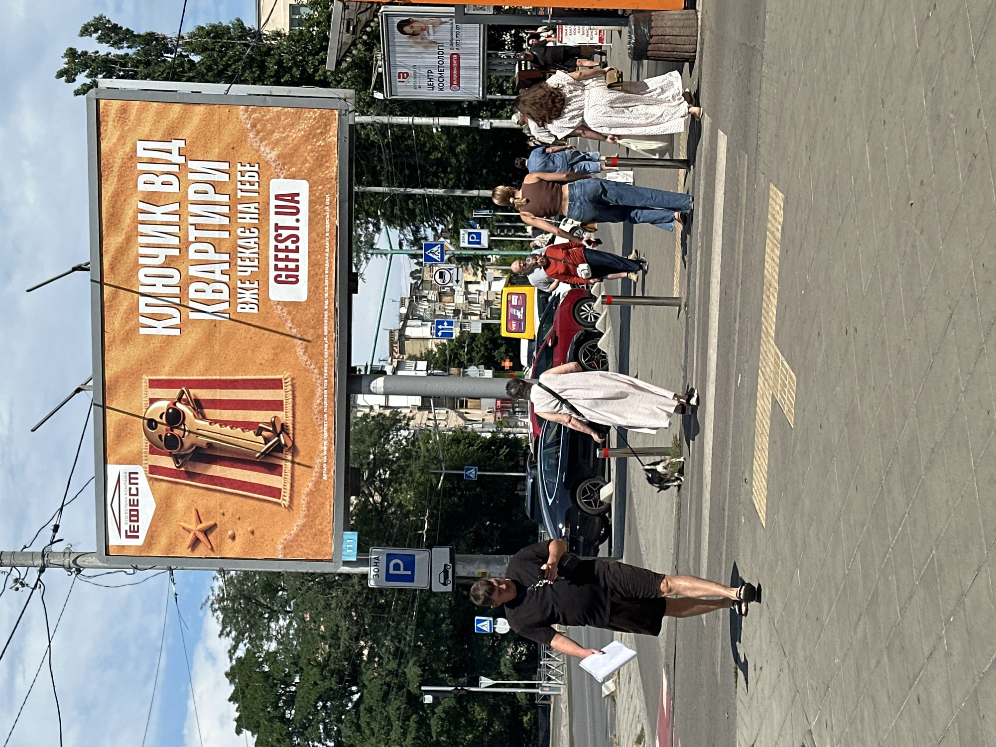 The image depicts a street scene in Odesa, Ukraine, on a sunny day. A large billboard advertises "GEFEST.UA" with an image of a key on a beach towel. In the foreground, a man in a black t-shirt and shorts walks on a sidewalk holding papers. He is looking at his phone. To his right, a woman in a white dress walks a small dog on a leash across a pedestrian crossing. Several other pedestrians are visible in the midground and background, along with cars and buildings. Traffic signs and street lights are also present. The overall impression is of an ordinary urban day in Odesa.
