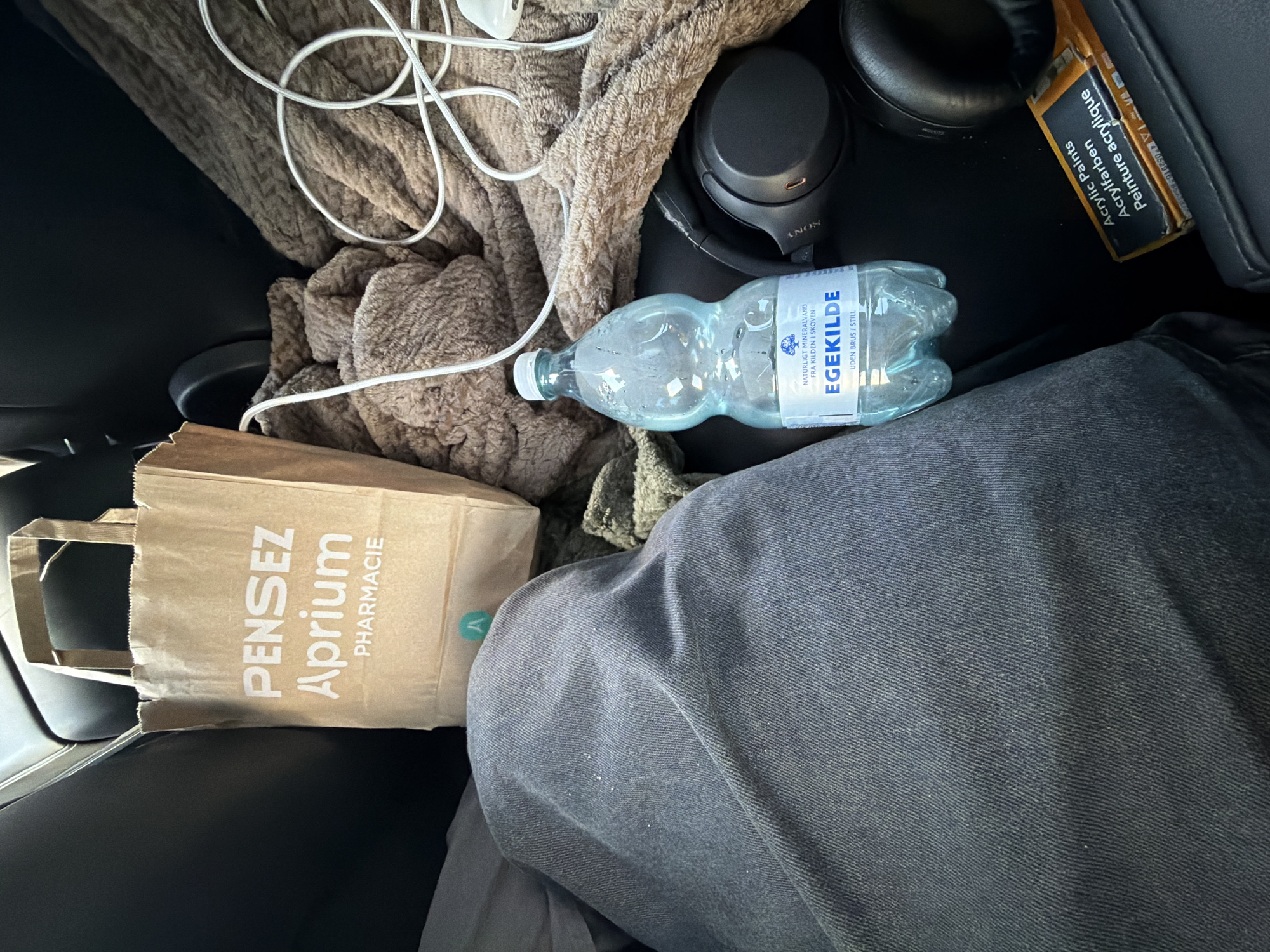 The image shows the interior of a vehicle. In the foreground, a person's dark denim-clad legs are visible. To the right of the legs, a brown paper bag with "PENSEZ Aprium PHARMACIE" printed in white text is situated. Next to the bag is a bottle of "EGEKILDE" natural mineral water. Further to the right, a pair of black Sony headphones rests on a brown, textured blanket. A white cord is also visible draped over the blanket. In the bottom right corner, a package labeled "Acrylic Paints Acrylfarben Peinture acrylique" is present. The overall scene suggests items belonging to a passenger in the vehicle. The location is identified as Ödsmål, Sweden.
