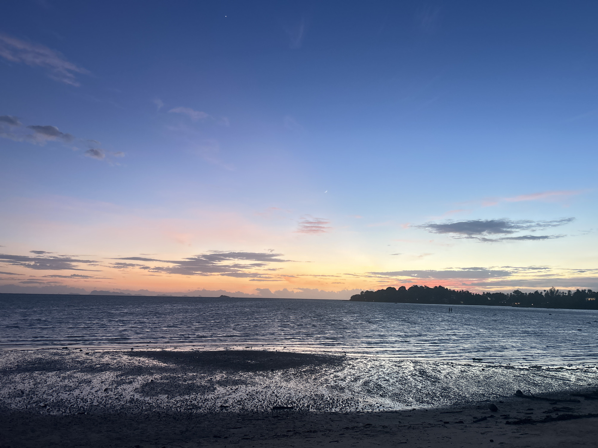 The image depicts a serene beach scene in the Ko Pha-ngan District of Thailand during twilight. The sky transitions from a deep blue at the top to lighter shades of pink and orange near the horizon, where clouds are scattered. A crescent moon is visible in the upper right portion of the sky. The sea is calm, with gentle ripples reflecting the colors of the sunset. The foreground is a sandy beach with areas of wet sand, likely exposed by the receding tide, creating a reflective, almost mirror-like surface. In the distance, a silhouetted treeline with scattered buildings marks the coastline. Two small figures, appearing to be people, are faintly visible standing in the shallow water in the middle distance. The overall atmosphere is peaceful and tranquil.