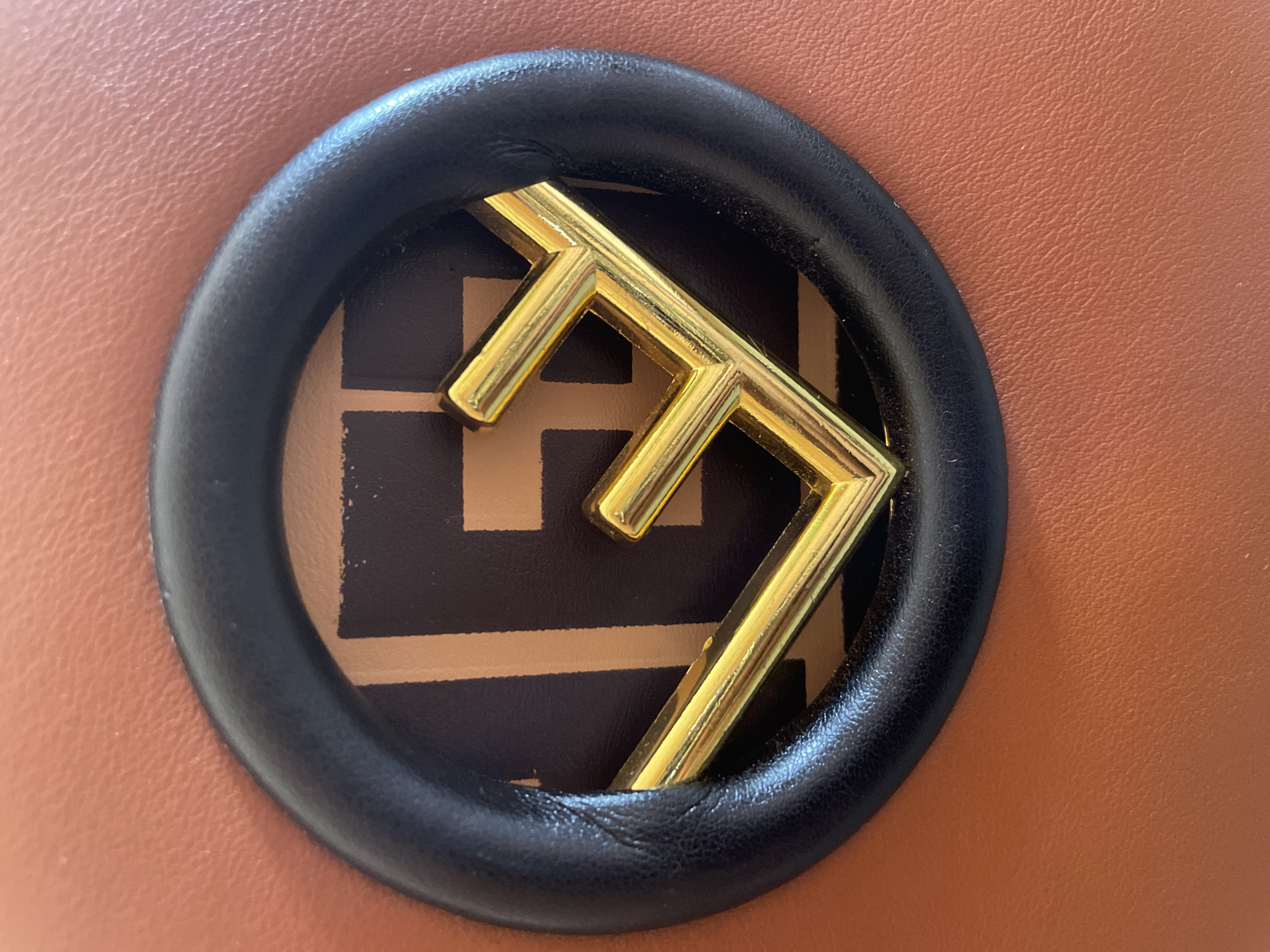 A close-up shot showcases the Fendi logo on a brown leather bag. The logo is gold-colored and features the iconic double 'F' design. It is set within a black leather circular frame. The texture of the brown leather is visible, appearing to be finely grained. The lighting suggests an indoor setting, possibly during the day, as there is a soft, even illumination across the surface. No people are visible in the image. The focus is entirely on the designer detail of the accessory. The background is the continuation of the brown leather material of the bag. There is no discernible text in the image other than the brand's logo.