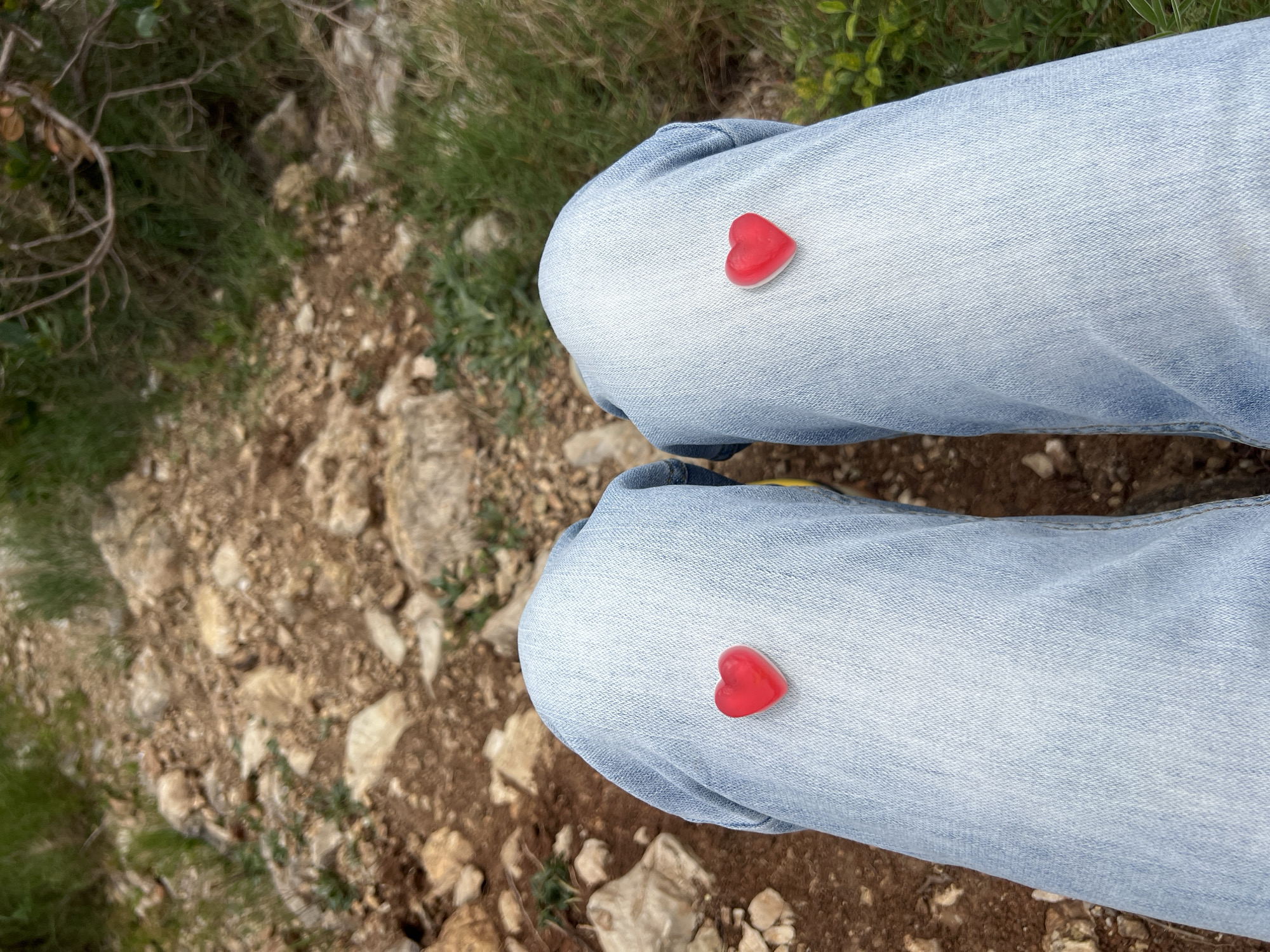 The image shows a view from above of a person's legs clad in light blue denim jeans. On each knee, there is a small, red, heart-shaped candy. The background consists of rough ground with patches of grass and small stones. The location is likely outdoors, possibly in a natural area near Budva, Montenegro, given the type of terrain. No specific landmarks or buildings visible to identify the exact location.