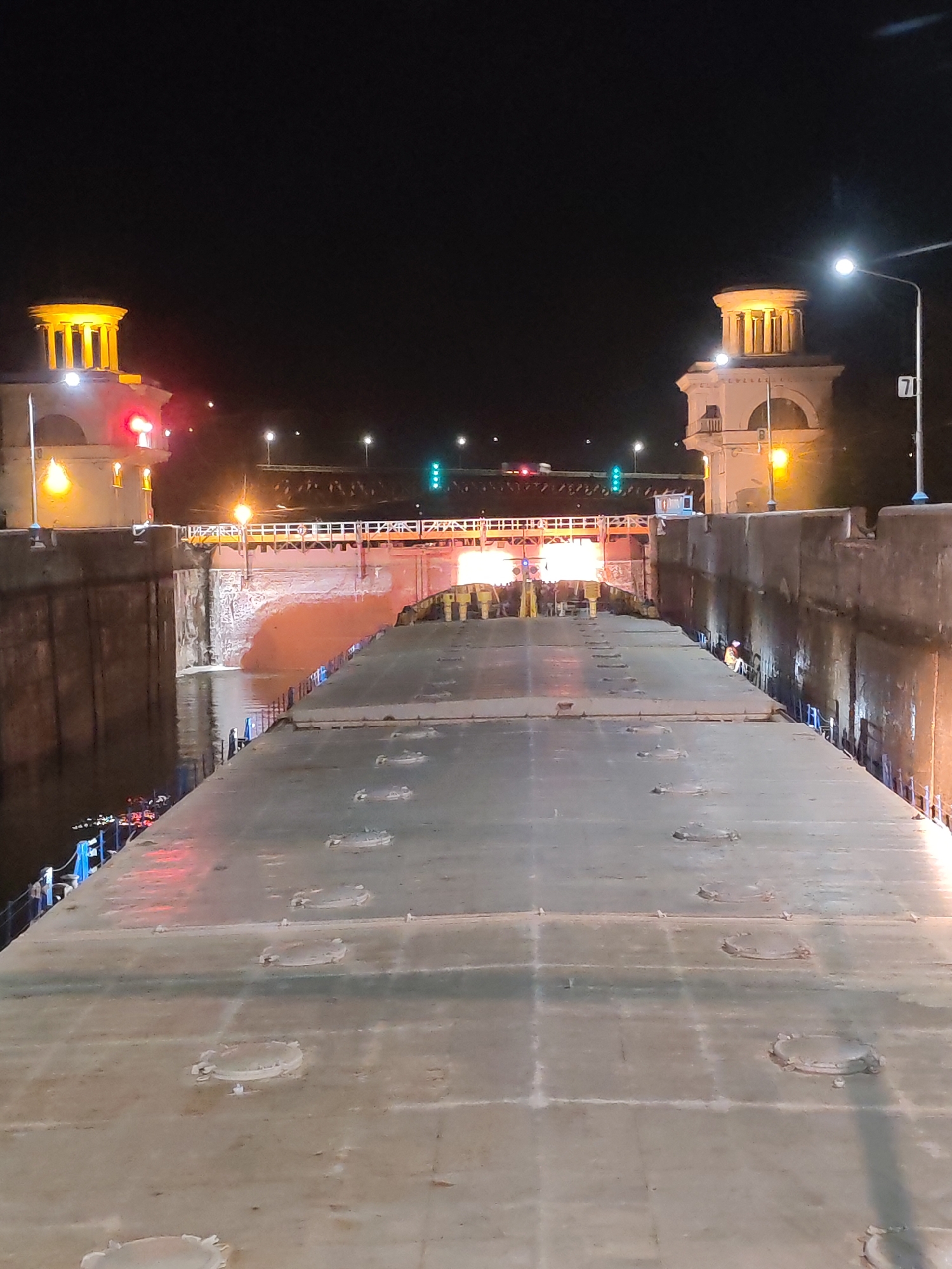 The image depicts a nighttime scene at a lock on the Volga River in the city of Ples, Russia. The view is from atop a barge or boat, looking forward through the lock. On either side are the walls of the lock, illuminated by lights, with two decorative towers at the far end. A pedestrian bridge crosses over the lock in the distance, with streetlights and traffic signals (red and green) visible beyond it. Water is present in the lock, reflecting some of the light. On the right, a sign post with the number 71 is visible. The topside of the barge is a flat, grey surface punctuated by numerous circular access hatches.