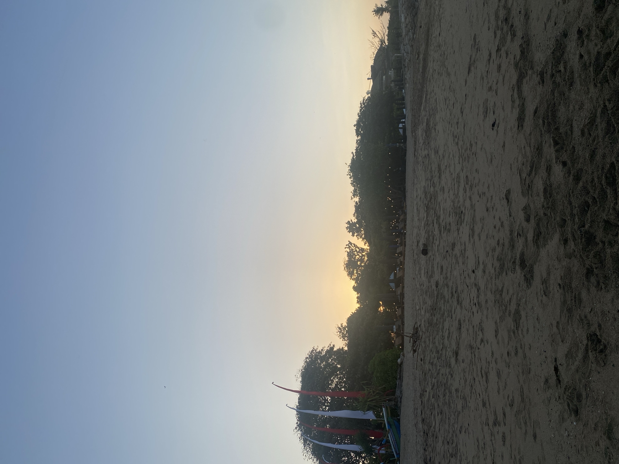 An outdoor, low-angle, full shot captures a beach in Denpasar, Indonesia, during dusk. The foreground is dominated by dark, textured sand with scattered footprints and a few pieces of driftwood. In the midground, a line of lush green trees forms a backdrop, with a subtle glow of sunset peeking through the foliage. Attached to the trees are several white and red flags on poles, and a small boat with blue and green accents rests on the sand. Further into the midground, beyond the trees, there are visible string lights, lounge chairs, and buildings with tiled roofs, suggesting a resort or restaurant area. The sky transitions from a pale yellow near the horizon to a soft blue overhead. A single bird is visible in the sky.