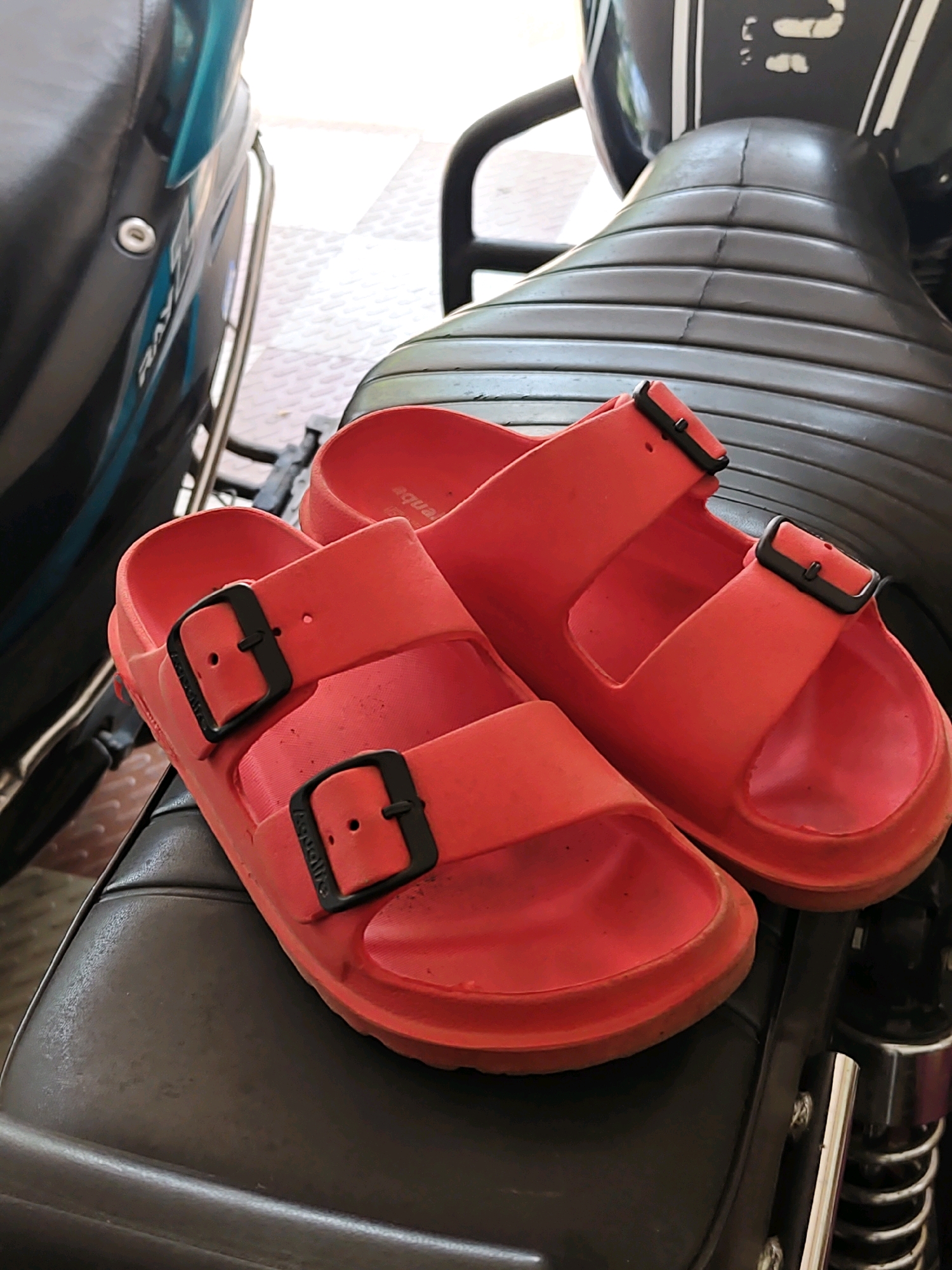 Two bright red slides with black buckles are placed on the dark brown, textured seat of a motorcycle. The slides are positioned diagonally, with one slightly overlapping the other. The left slide is in the foreground and is closer to the viewer, while the right slide is partially obscured by the left one. The motorcycle seat appears worn with visible creases and possibly some dirt. In the background, a portion of a motorcycle's teal-colored bodywork is visible on the left, and a lighter-colored tiled surface is on the far right. The scene is outdoors, with natural lighting. The location is in Pukkattupady, India.