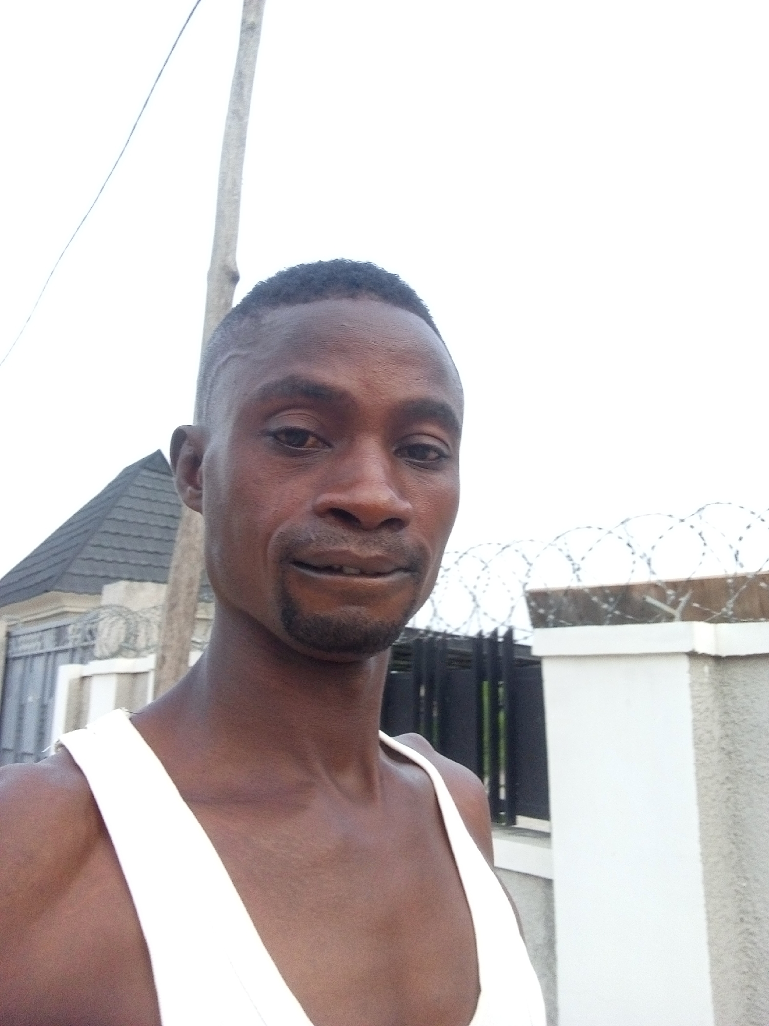 A medium shot captures a dark-skinned man with short-cropped, dark hair and a trimmed goatee. He is wearing a white tank top.
Behind him, in the background, is a fence topped with coiled razor wire. Adjacent to the fence is a building with a dark grey roof. A utility pole stands beside the building. The sky overhead is overcast.
Given the prompt's request to locate the image in Adonkolo, Nigeria, and the presence of fences with razor wire, the location may be a residential area or a secured compound within the city. The specifics are challenging to determine with the limited visual context.