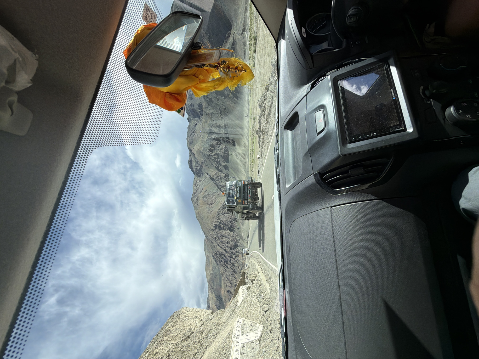 The image is taken from the interior of a vehicle looking out through the windshield at a mountainous landscape. A dark-colored military truck is driving on a paved road ahead, with a white vehicle following it. The road is flanked by rocky, barren hills under a partly cloudy sky. Inside the vehicle, the dashboard, a rearview mirror with a yellow and orange religious offering hanging from it, and a portion of the steering wheel are visible. The scene appears to be located in Chumathang, India, a region known for its rugged terrain and military presence.