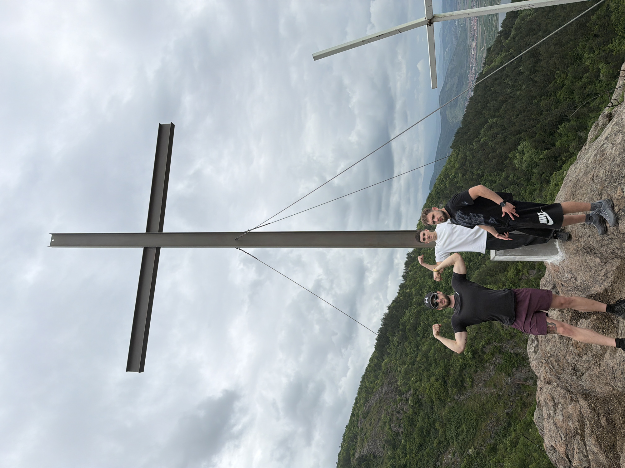 Three men are standing on a rocky outcrop near two metal crosses, one larger and made of I-beams and the other smaller and tubular. The scene is at an elevated location overlooking a green valley, likely near Ustina, Bulgaria. The sky is cloudy. The man closest to the camera is wearing a black T-shirt, maroon shorts, sunglasses, and a black baseball cap, and is flexing his biceps. The middle man is wearing a white T-shirt. The man furthest from the camera is wearing a black T-shirt and black Nike shorts. All three men are standing near the lower portion of the larger I-beam cross. The rocky outcrop leads down to a forested hillside. Beyond the hillside, the valley opens up to a town. A second, smaller, tubular cross is visible at the very top of the frame. Wires can be seen connecting the crosses.