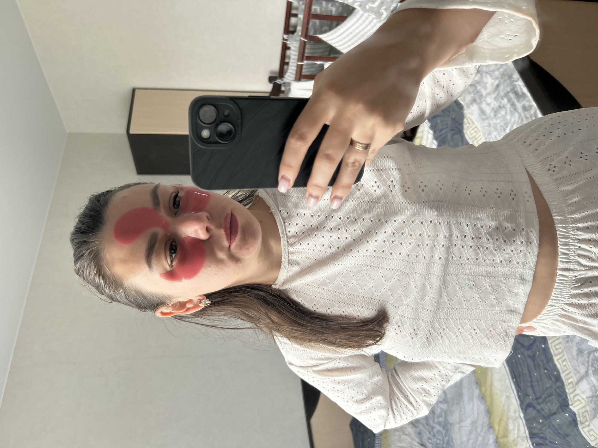 A woman with dark hair pulled back is taking a selfie in a bedroom. She is wearing a white, eyelet top and white shorts. Red under-eye masks are applied to her face. She is holding a black smartphone in her right hand. The background shows a bed with patterned bedding, a closet, and a crib. The lighting suggests it is daytime. The user specified that this image is located in Chaikovsky, Russia.