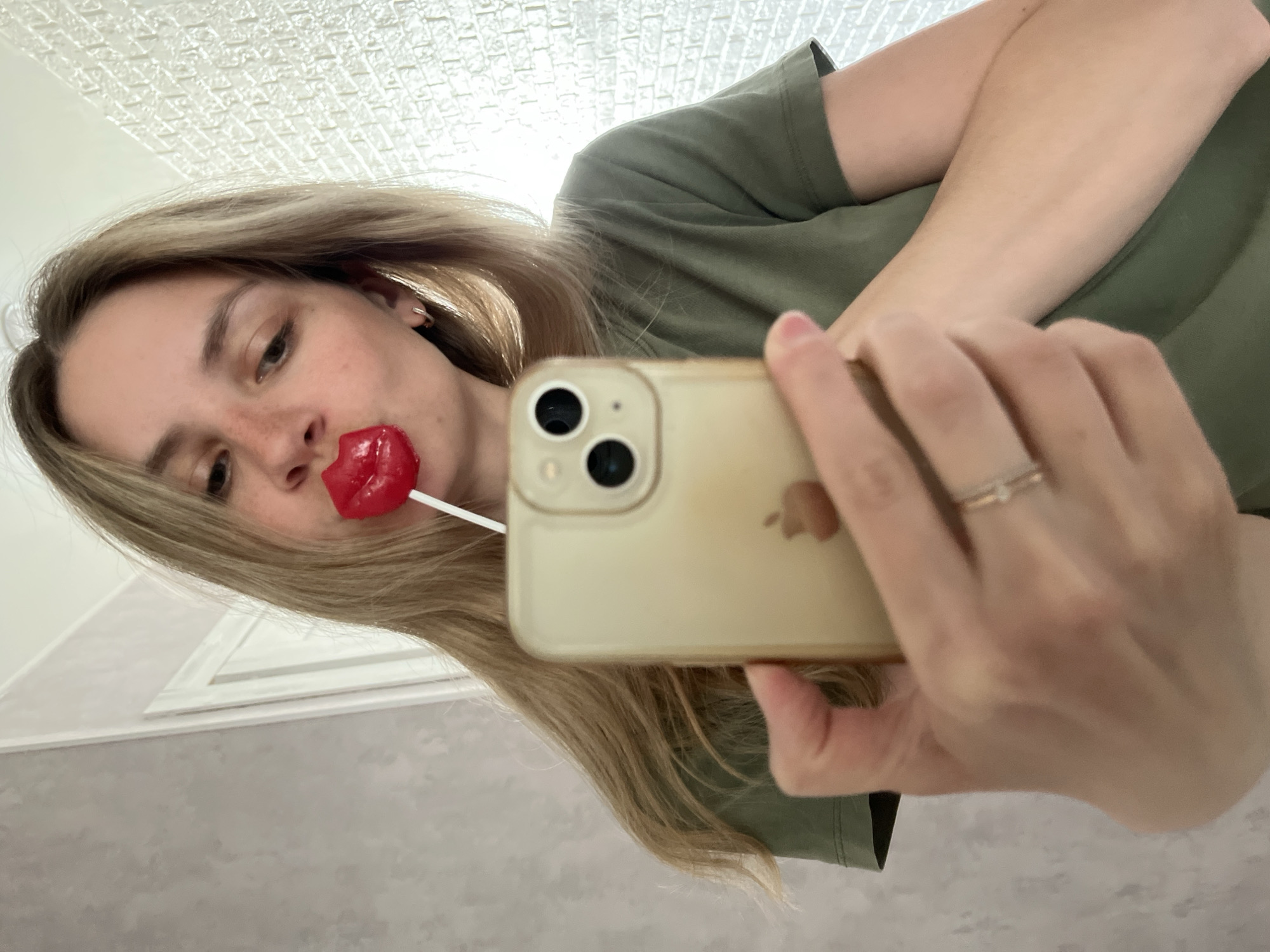 A young woman holds an iPhone in her right hand, angled towards her face, capturing a selfie. She is wearing a light green t-shirt and has a red lip-shaped lollipop in her mouth. Her hair is blonde. She appears to be wearing a ring on her right ring finger. The background includes a textured white ceiling and a section of a grey and white wall. The image does not provide sufficient information to confirm the location as Chaikovsky, Russia.