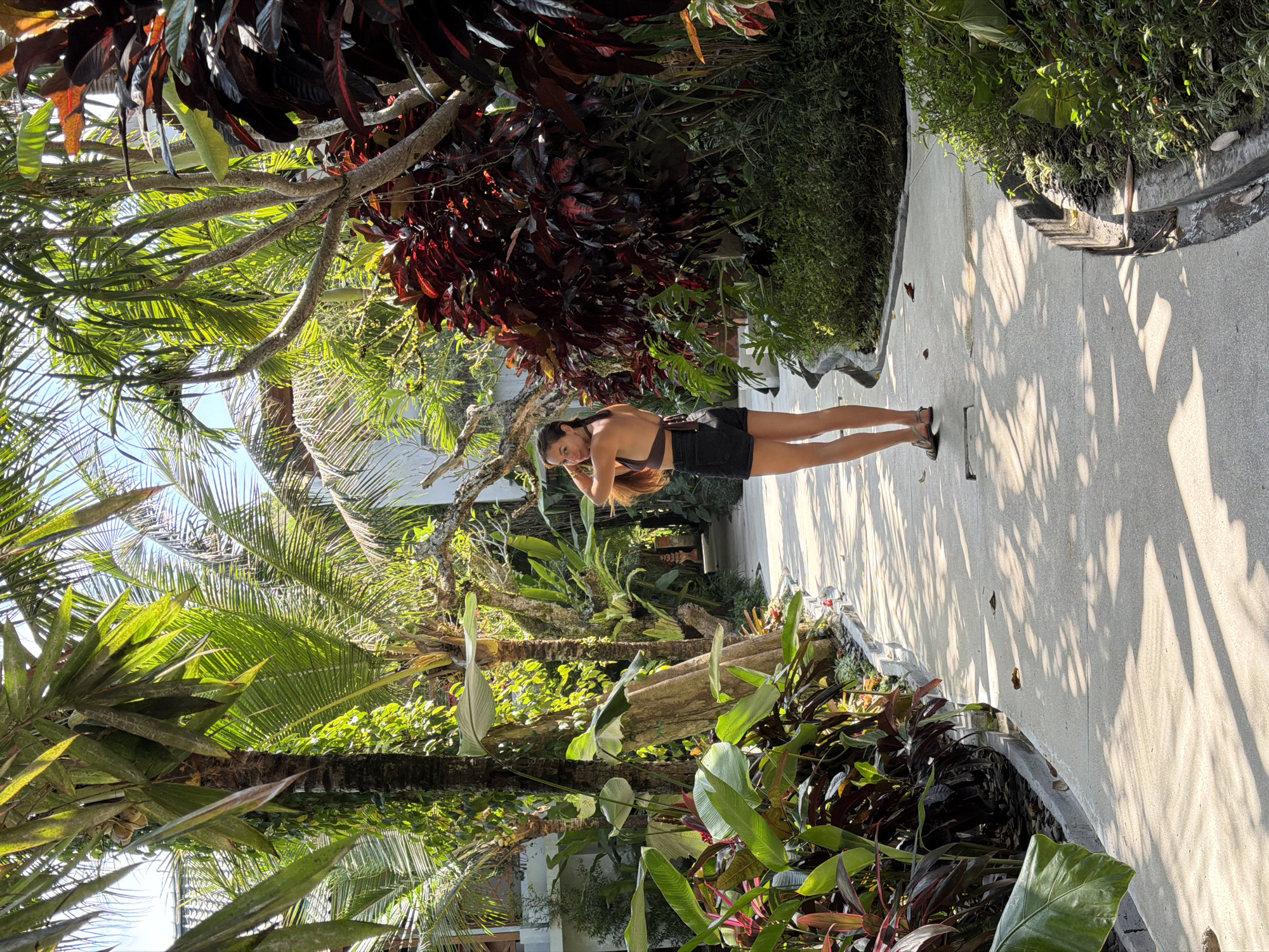 A woman stands on a paved pathway in a lush, tropical garden environment, likely in Ubud District, Indonesia. The pathway is lined with dense foliage, including palm trees with fronds casting dappled shadows across the ground, and large-leafed plants with red and green coloration. The woman is wearing a dark brown halter top and black shorts, with her hair partially pulled up. She is facing away from the camera, looking over her shoulder with one hand near her head. The scene is brightly lit by sunlight filtering through the trees.