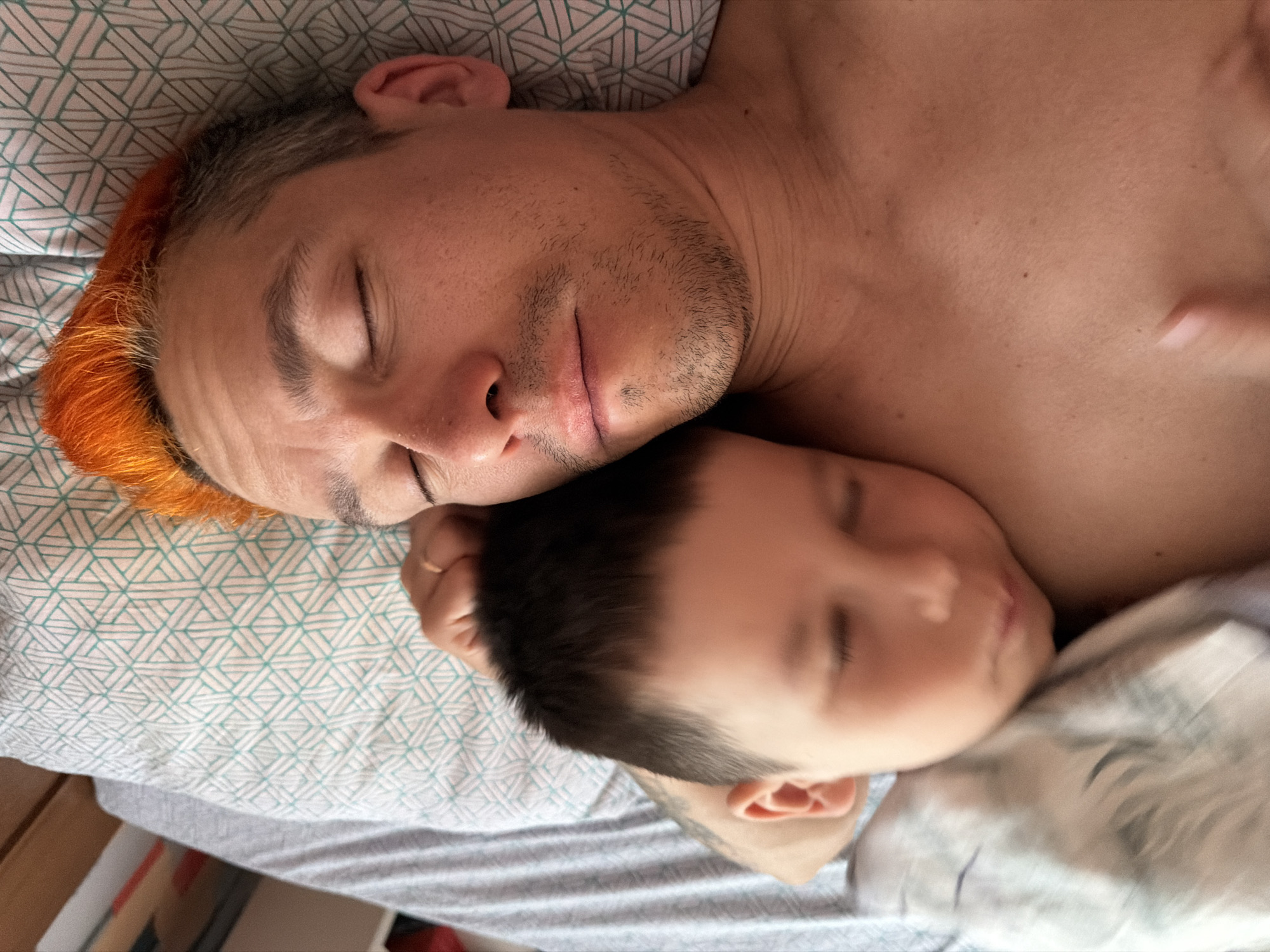 A man and a child are lying down on a bed, possibly sleeping. The man is fair-skinned with some facial hair and orange hair. The child is fair-skinned with dark hair. They are lying on a pillow and a bed with patterned bedsheets. The image location is possibly in Chaikovsky, Russia.