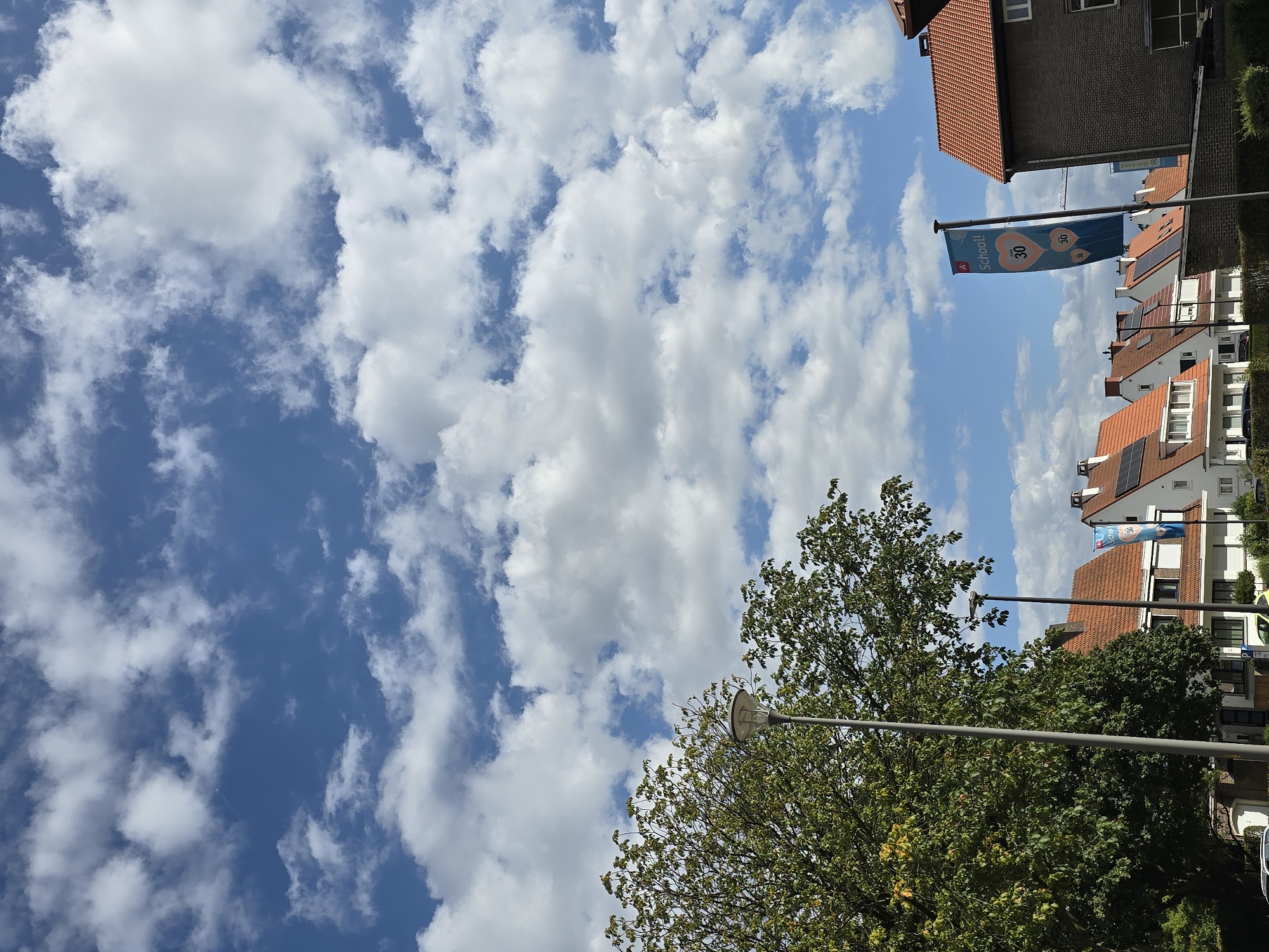 The image depicts a street scene in the city of Antwerp, Belgium, under a partly cloudy blue sky. In the foreground, a large tree with green leaves dominates the left side of the frame, partially obscuring a grey lamppost. To the right of the tree, a series of residential buildings with terracotta tiled roofs are visible. Some of these buildings have solar panels installed on their roofs. A blue banner with the word "School!" and heart-shaped symbols with the number "30" on them is flying from a flagpole in front of one of the buildings. The background shows more buildings and a bright, expansive sky filled with scattered white clouds. The scene appears to be during daylight hours, possibly late morning or early afternoon, given the lighting conditions. There are no people or vehicles clearly visible in the image.