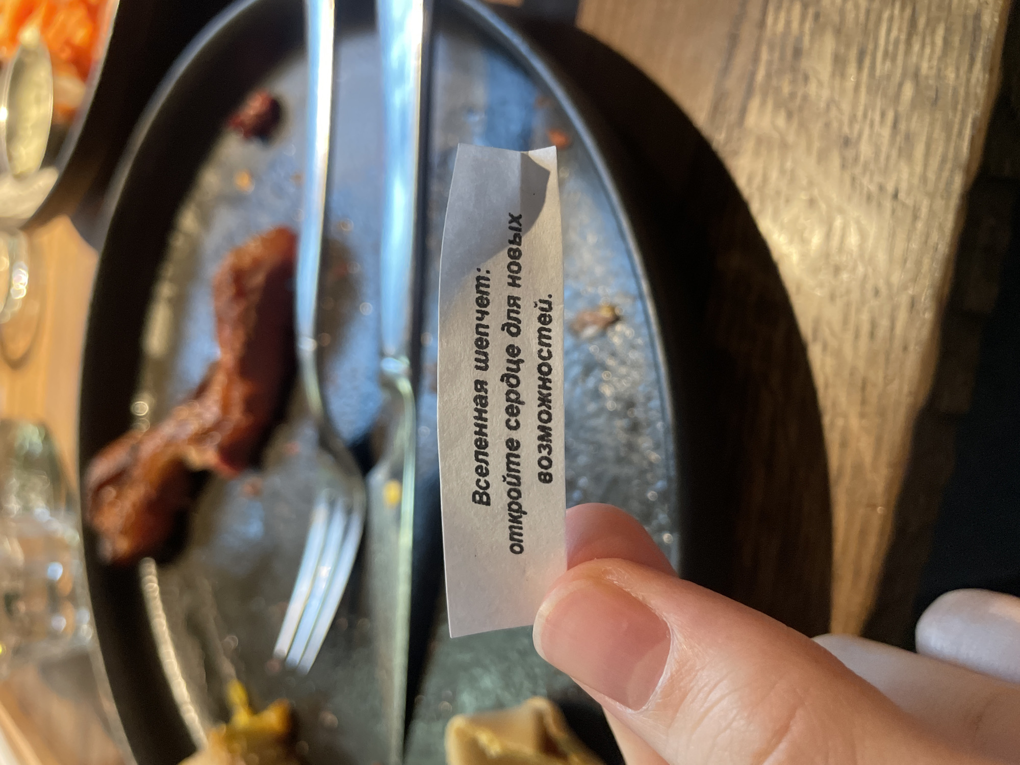 A hand holds a fortune cookie slip with text written in Russian: "Вселенная шепчет: откройте сердце для новых возможностей" ("The universe whispers: open your heart to new opportunities"). The hand is positioned over a dark, partially eaten meal with a fork and knife laid on top. The photo was likely taken at a table in a restaurant. Location cannot be determined from the image.