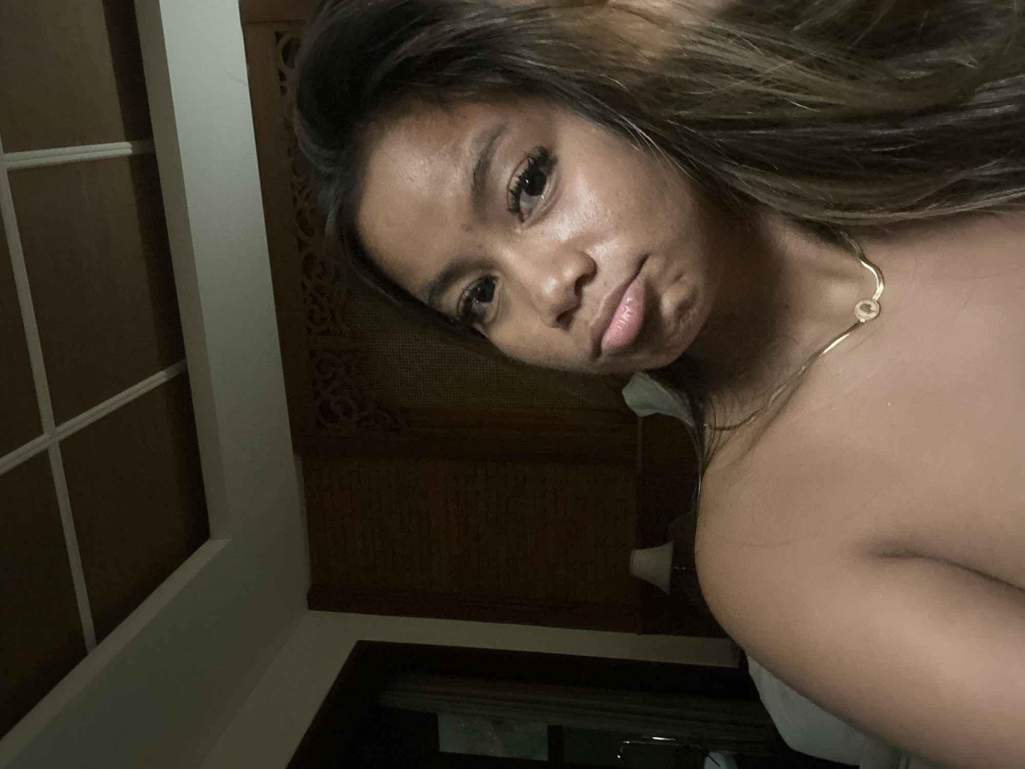 A young woman is shown in a close-up shot. Her skin is tan and she has long, dark hair with blonde highlights. She is wearing a gold necklace. She is looking at the camera with a slight pout. The background includes a window with white panes, a dark wooden door or wall with an intricate pattern, and a lamp. The setting appears to be indoors, possibly a hotel room or residence. The location is likely in Ubud District, Indonesia, based on the prompt's information.