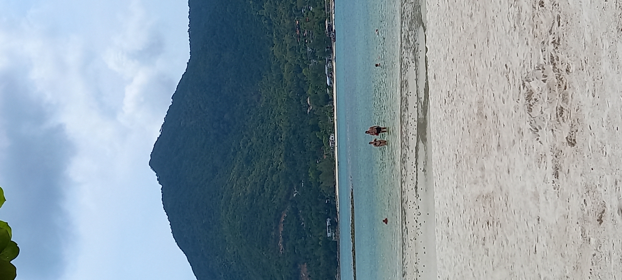 The image captures a beach scene in Baan Chaloklum, Thailand. In the midground, two individuals are wading in the shallow, clear water near the shoreline. Further out in the water, a single person is visible. The beach itself is a light-colored sand, marked with tidal patterns. To the left, a large, lush, green mountain occupies a significant portion of the frame, stretching upwards towards a sky that is partially obscured by clouds. Buildings are sparsely visible along the base of the mountain, indicating some development near the coastline.