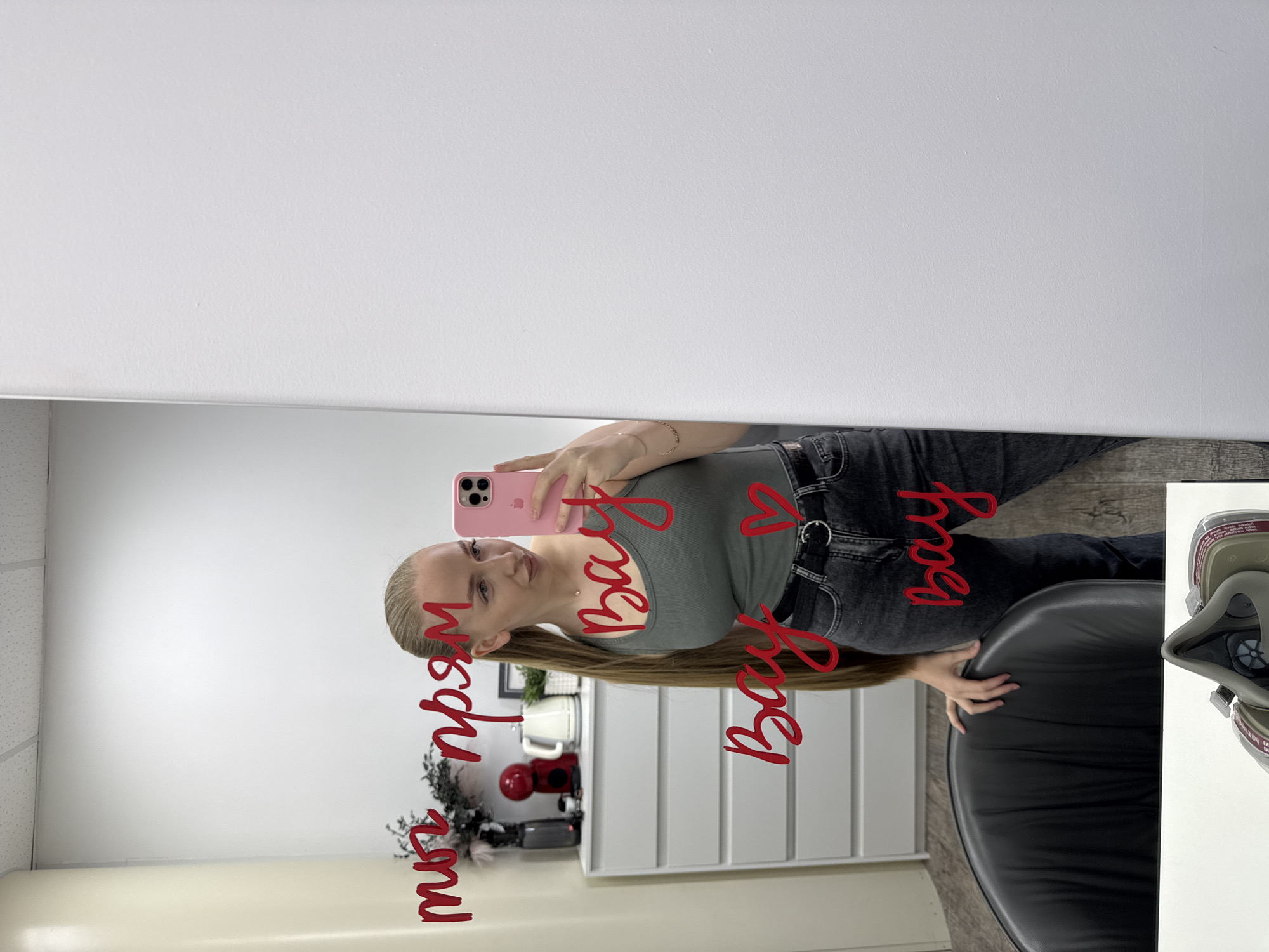 The image depicts an indoor scene, likely a commercial or private interior space, that does not contain identifiable features to confirm the specified location of Petropavlovsk-Kamchatsky, Russia. The central subject is a young adult female, reflected in a large rectangular mirror, engaged in taking a selfie with a pink smartphone (likely an iPhone 13 Pro/Pro Max or 14 Pro/Pro Max).
The woman has long, straight, light-colored hair, styled in a high ponytail that extends significantly down her back. She is wearing a dark olive green or grey tank top and dark-wash jeans secured with a black belt featuring a circular silver-colored buckle. A thin, gold-colored bracelet is visible on her left wrist. Her left hand rests on the back of a dark-colored, possibly black, upholstered chair, visible in the lower left portion of the reflection. She is looking towards her phone, held in her right hand, with a slight expression.
Superimposed in red script on the mirror's surface are the words "ты прям Вау Вау Вау" (translating to "You are just Wow Wow Wow" or "You are really Wow Wow Wow"), followed by a small red heart symbol.
The background visible in the mirror's reflection shows plain white walls and a white acoustic tile ceiling. To the left of the woman, a white chest of drawers or dresser with multiple horizontal panels is positioned against the wall. On top of this furniture are several items: a tall, dark vase containing dark feathery or leafy decorative branches, a bright red compact coffee machine or appliance, a white electric kettle, and a small black-framed object, possibly a picture frame. The floor visible in the reflection is a light wood-effect laminate or tile.
At the very bottom center of the original image, outside the mirror's reflection, a partial view of a grey-green respirator mask with visible filters is present, suggesting its placement on the floor or a low surface.
