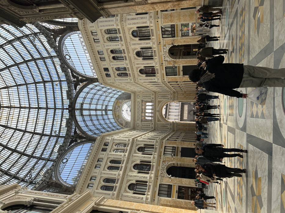 The Galleria Umberto I is a 19th-century shopping arcade located in the heart of Naples, Italy, at the coordinates 40.839° N, 14.248° E. The Galleria, adorned with a large glass and iron dome, is comprised of two intersecting arcades lined with shops and restaurants. The image captures a bustling scene with a mix of people, some shopping, others strolling, and still others sitting in cafes. The marble floor is patterned with geometric shapes and inlaid with mosaics. The bright sunlight streams through the glass dome, illuminating the arcade. There is a large open area in the center of the Galleria, surrounded by shops and restaurants. The back wall of the Galleria features a large arched doorway leading out to the city. The image was taken during the day, possibly on a weekday, as there is a considerable number of people moving about the arcade.