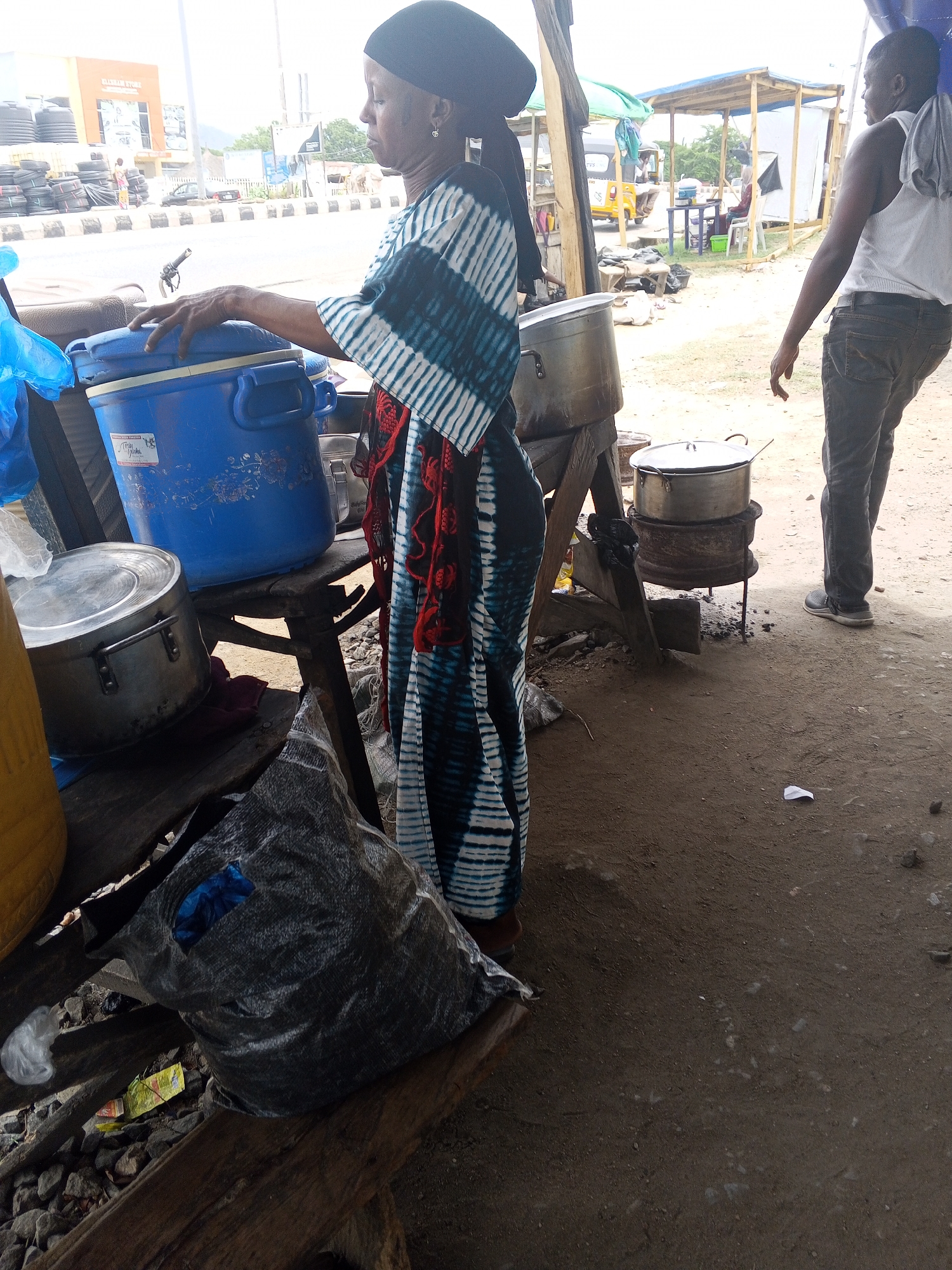 In Lokoja, Nigeria, a woman wearing a black head covering and a long, patterned dress stands next to a table holding a blue container and other cooking equipment. A man stands nearby, and in the background, a building, vehicles, and a simple shelter are visible. Cooking pots rest on a stove made from a metal tire. The ground appears to be dirt or gravel. A bag is on the ground under the table.