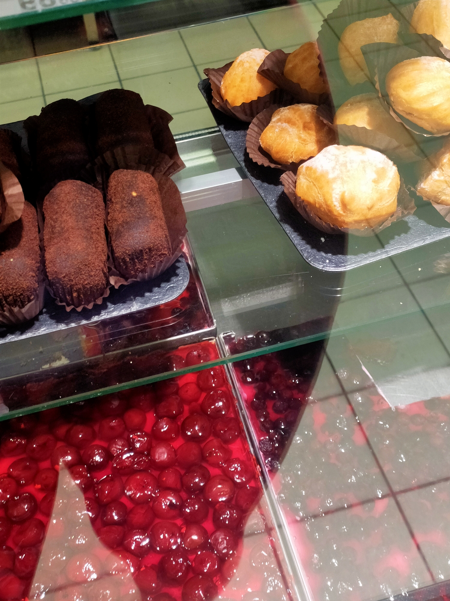 The image displays a selection of pastries in a refrigerated display case, likely in a bakery or cafe. The subjects are various sweet treats: several chocolate-covered, cylindrical pastries dusted with cocoa powder are visible in the foreground on the left, presented in dark brown paper wrappers. To the right, a selection of golden-brown, cream-filled pastries, possibly eclairs or profiteroles, are arranged in dark paper cups on a slate tray. In the lower portion of the image, a vibrant red dessert filled with whole cherries is visible, beneath the pastries, suggesting a layered cake or tart.
The setting appears to be indoors, with the bright lighting and reflective glass surfaces indicating a commercial establishment. The overall impression is one of a well-stocked pastry counter, inviting customers to choose from a variety of desserts. The arrangement and presentation of the pastries suggest they are ready for sale. There are no people visible in the image, and no specific time of day or weather conditions can be inferred. No visible text is present. The location context is Kyiv, Ukraine, which implies a European culinary style for the pastries.