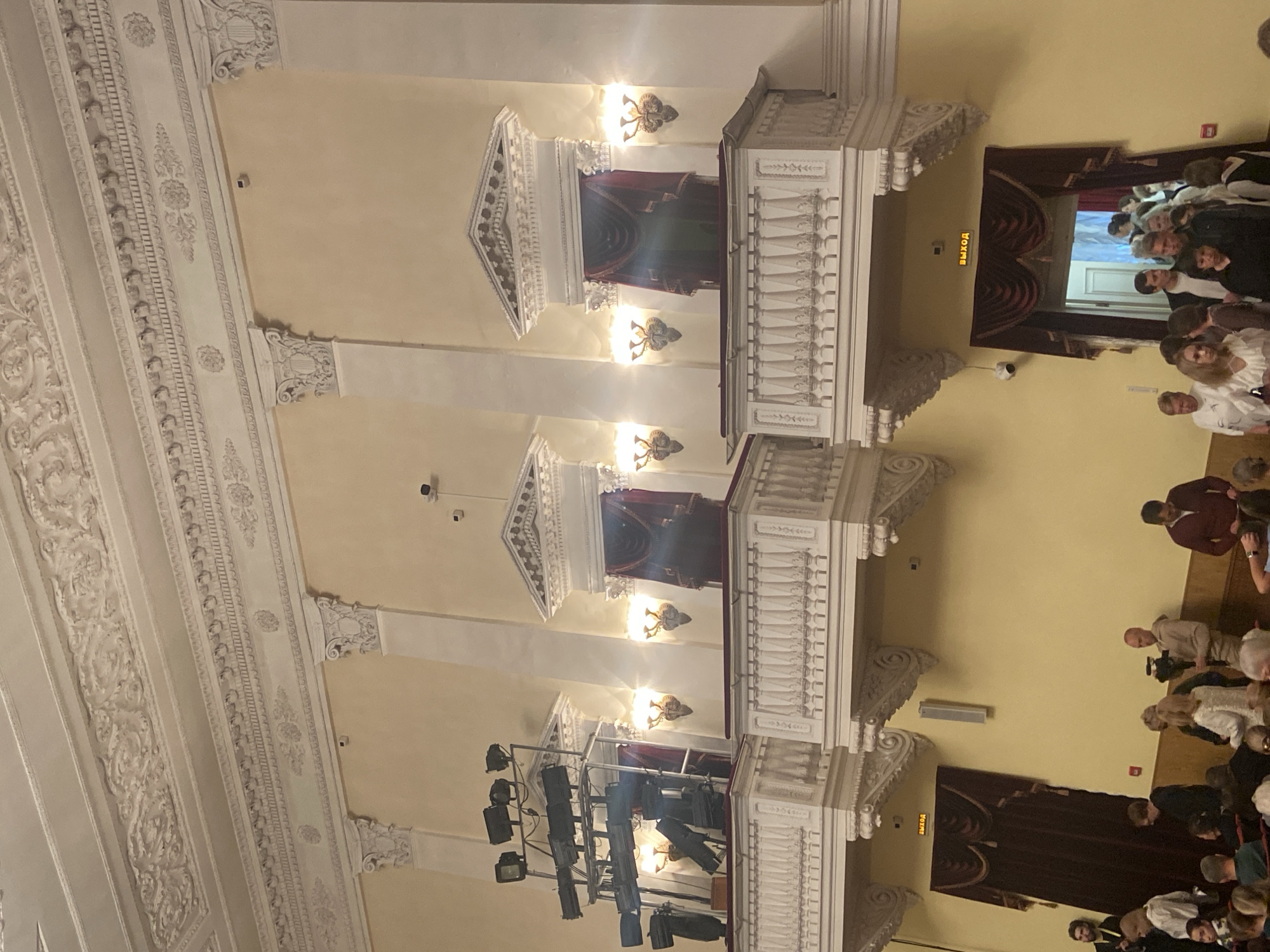 The image depicts the interior of a theater or concert hall. There are three balconies on one side, ornately decorated with white railings and classical architectural details. The walls are painted in a pale yellow hue, and the ceiling is adorned with elaborate white molding. A lighting rig is set up below the lowest balcony. People are gathered in the auditorium, with most facing away from the camera. A doorway with a red curtain is visible at the end, bearing the word "выход" which means "exit" in Russian.
While the image does not contain specific clues to confirm its exact location in Sergiyev Posad, Russia, the architectural style is consistent with theaters from the 19th or early 20th centuries, which can be found in many cities in Russia. Without further clues it is not possible to establish location certainty.