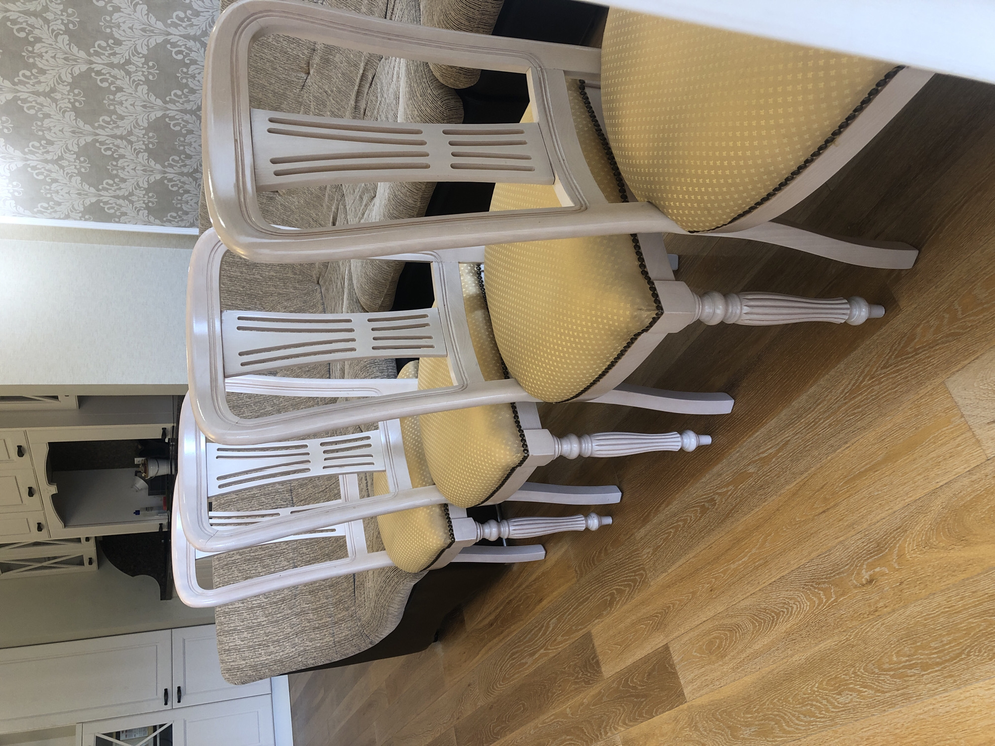 The image shows a room interior featuring four chairs. The chairs have a similar design with a cream-colored wooden frame and yellow patterned seats. The chairs are arranged in a row on a wooden floor. In the background, a section of a beige sofa is visible, along with a wall decorated with patterned wallpaper. Part of a white kitchen cabinet is also seen in the background on the left. The room appears to be well-lit. There is no indication that this image was taken in the city of Chaikovsky, Russia.