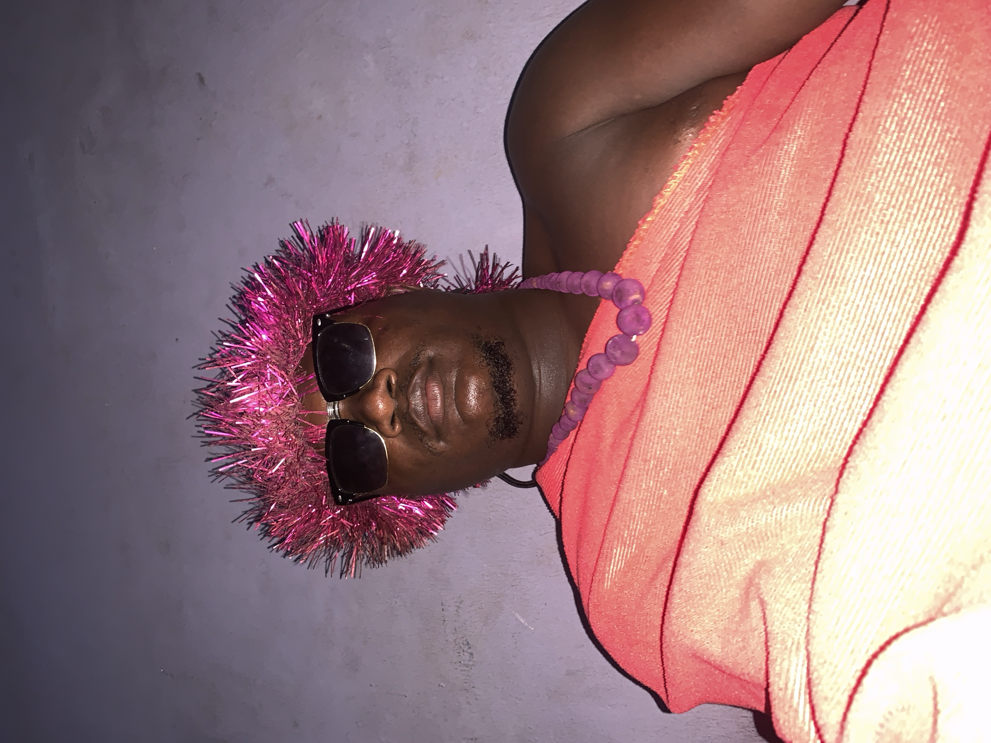 A man, presumably of West African descent, is pictured lying down and looking at the camera. He is wearing a pink, spiky wig, dark sunglasses, and a purple beaded necklace over an orange draped garment that covers his torso and shoulder. The background is a plain, textured, grayish wall. The image quality is slightly grainy, and the lighting appears to be artificial, casting shadows on the wall. The overall impression is that of a celebratory or costume-related occasion, possibly taken indoors or in a dimly lit outdoor setting within Akwanga, Nigeria.