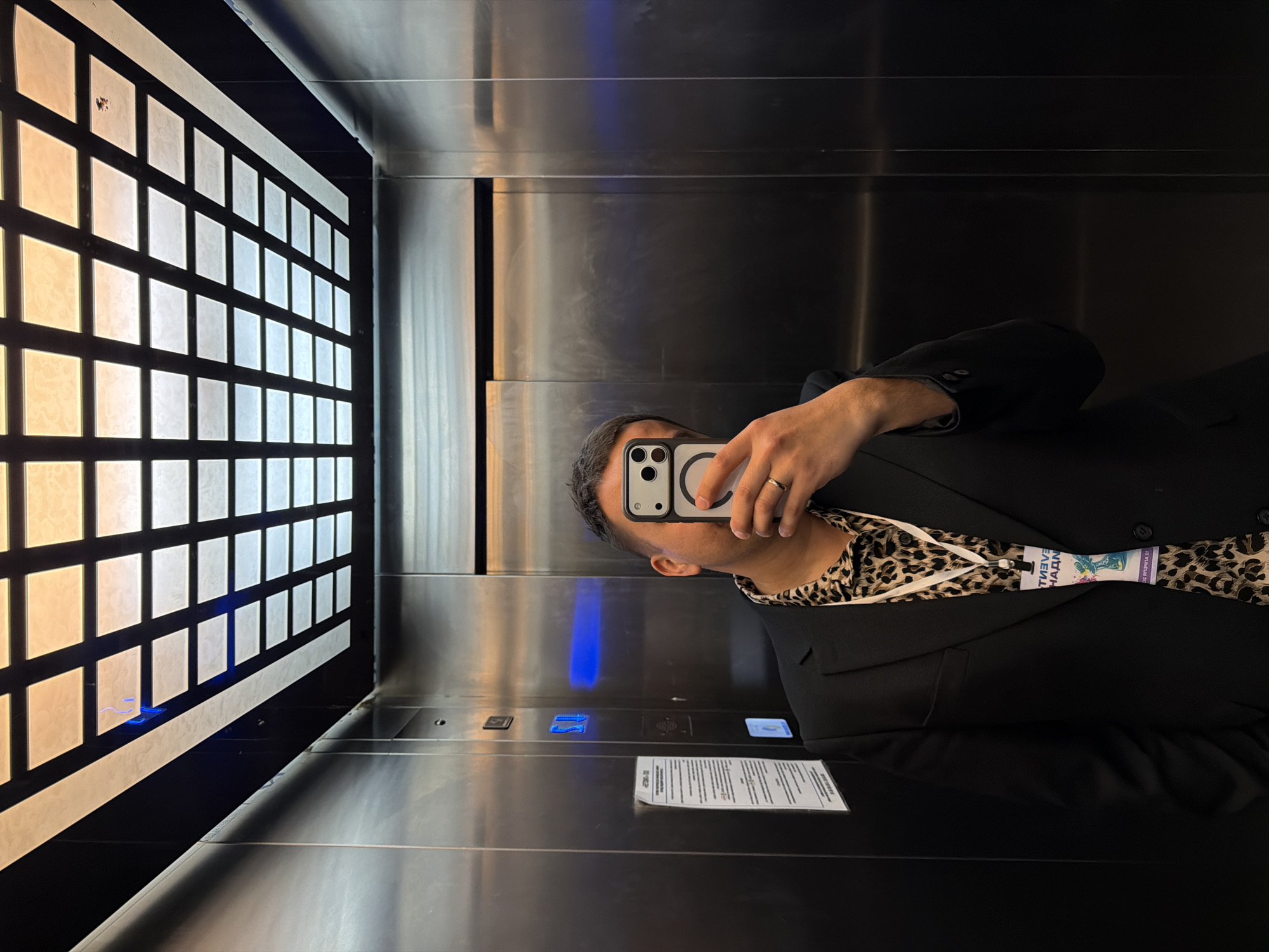 A person is taking a selfie in an elevator with a grid of illuminated panels on the left wall. The person is wearing a black blazer over a leopard-print shirt and has a lanyard with a name tag. Elevator buttons and information are visible on the mirrored metal wall. A reflection suggests the location is in Perm, Russia, based on writing on the name tag.