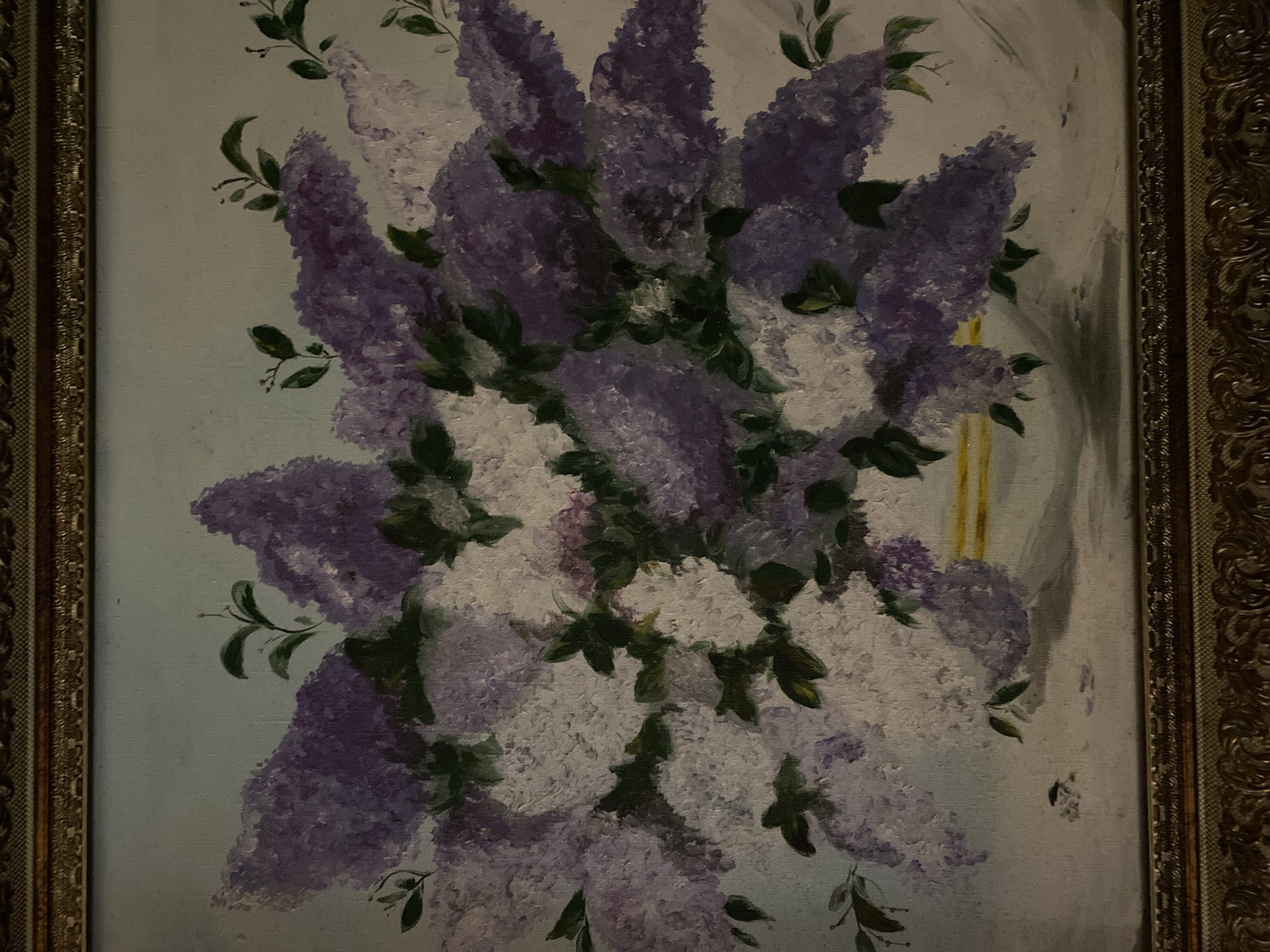 This is a close-up, angled, indoor photograph of a painting. The painting depicts a bouquet of lilacs, with both purple and white blooms, arranged in a vase. The vase appears to have a yellow band around its rim. The background of the painting is a pale, desaturated blue. The painting is framed by an ornate, gold-colored frame. The image does not provide any information about the city of Poltava, Ukraine, or any specific location within it. The photograph is of a static artwork and does not depict any people, actions, or interactions.