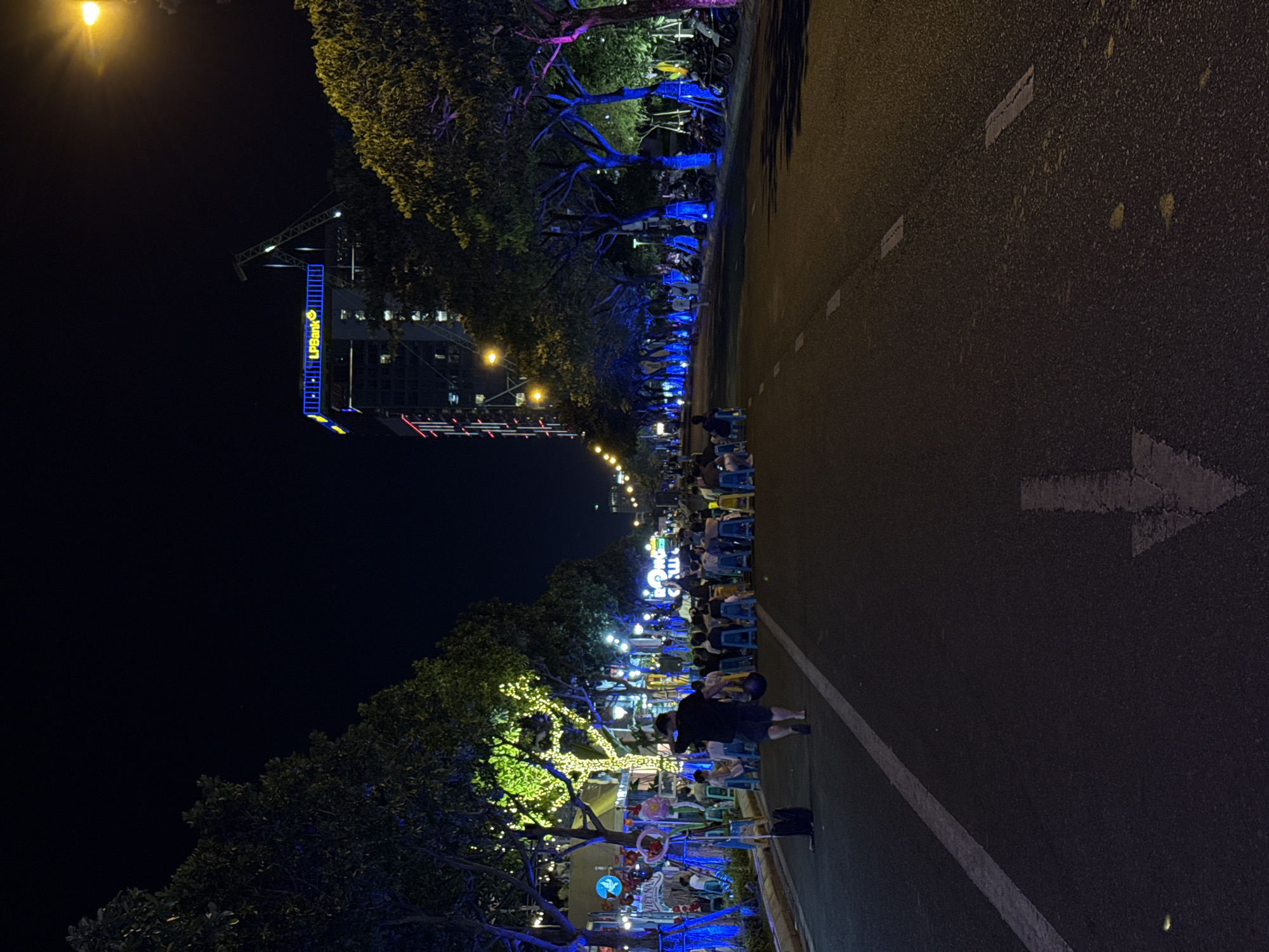 This nighttime image depicts an urban street scene in Đà Nẵng, Vietnam. The perspective is from the center of an asphalt road looking forward, illuminated by artificial lighting.
In the foreground, the dark asphalt road features a solid white line marking the left edge and a broken white line further right. A large, worn white arrow pointing away from the viewer is painted on the road's right side. A person, wearing a dark t-shirt and dark shorts, walks barefoot on the left side of the road, moving away from the camera. They carry two dark, bowl-shaped objects in their left hand. A black bag rests on the road to their immediate left.
On the left side of the street, a row of trees lines a sidewalk. The lower trunks and branches of these trees are wrapped in bright blue LED lights. Further down the row, one prominent tree is densely covered in small, glowing yellow-green fairy lights. Behind these trees, a gathering of individuals is observed, with many people seated on small blue plastic stools. A sign partially displaying "VIỆT NAM" is visible among the trees and seating areas.
On the right side of the street, another row of trees is present, illuminated with blue and some pink lights. Numerous motorcycles are parked along the curb under these trees. People are also visible walking and standing on this side, some seated on similar blue stools.
In the background, a tall building stands prominently on the right. Its rooftop features an illuminated sign displaying "LPBank" in yellow text on a blue background. Red vertical light strips are visible on the building's facade. A large construction crane is silhouetted against the dark sky behind the building. The sky is dark, indicating night, with no stars visible. Distant city lights extend down the street.