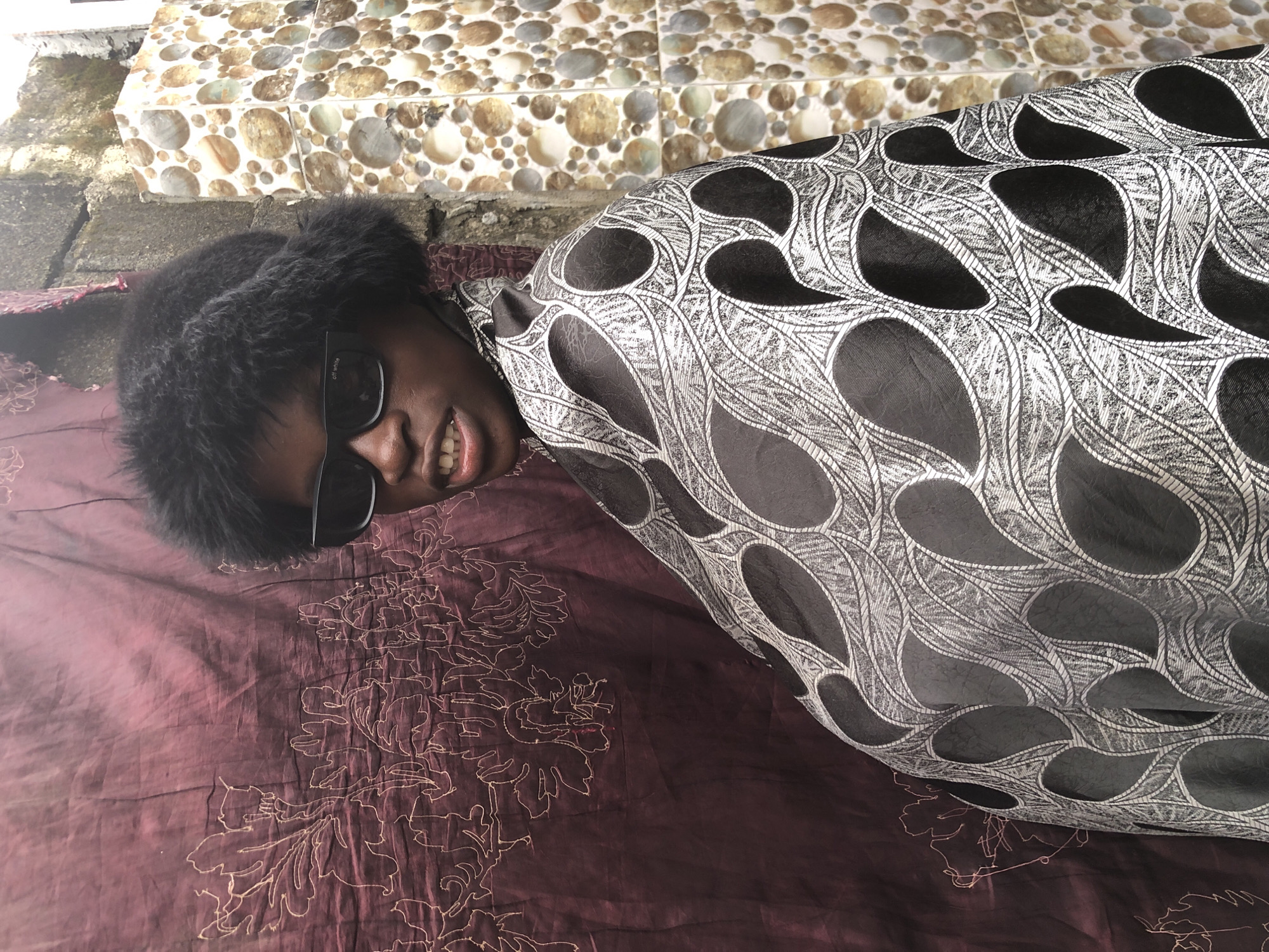 A person wearing a black fluffy hat and black sunglasses is pictured in an outdoor urban setting in Ado, Nigeria. The person is draped in a black and silver patterned fabric, which drapes over their shoulders and chest. Behind the person, a section of a maroon fabric with a white floral pattern is visible on the left. To the right, a wall covered in small, round, multi-colored tiles serves as a backdrop. The person's mouth is slightly open, revealing their teeth.