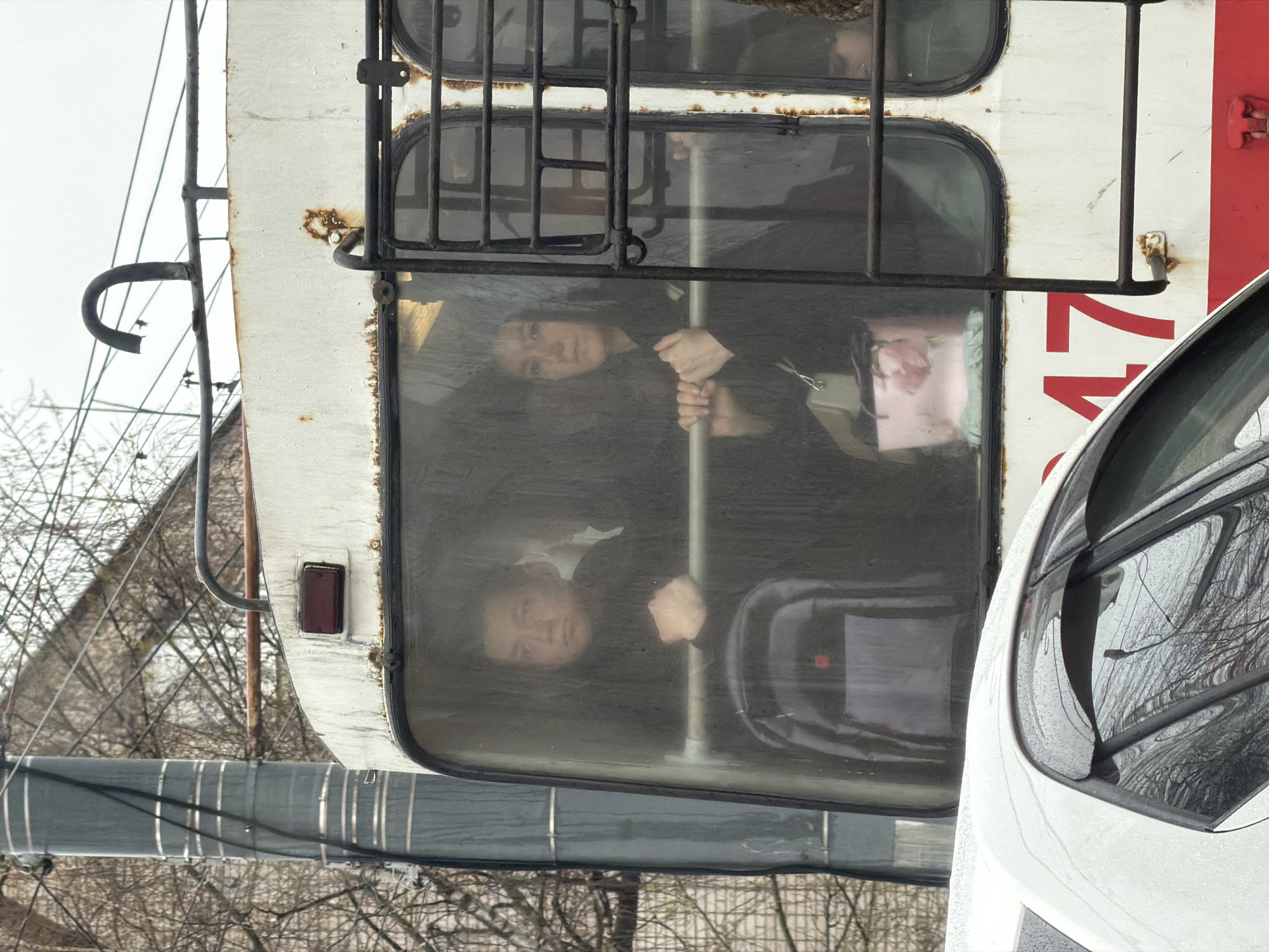 This image depicts a street scene, likely in Izhevsk, Russia, featuring a large public transport vehicle (possibly a trolleybus or bus) on the left and a white car partially visible on the right. The image is taken from a slightly elevated or angled perspective, looking into the windows of the public transport vehicle.
The large vehicle appears worn, with white siding, red markings on the right side, and visible rust around its window frames and metal fixtures. A protective metal grille covers some of its windows. Inside, two people are clearly visible through the glass. A woman, seemingly young, is in the upper part of the window, holding onto a vertical pole and looking directly towards the camera. Below her, another individual, a man, is also visible, resting his hands on a bar or surface and looking out. Both appear to be passengers. A pink object, possibly a bag or accessory, is seen near the woman.
On the far right, part of a modern white car is visible, reflecting the bare trees of the background in its windows.
The background consists of a light, overcast sky and a network of bare tree branches, suggesting a late autumn, winter, or early spring season. Overhead wires and utility poles are also present, indicative of an urban or suburban setting. A thick, grey cable or pipe runs horizontally in the lower left portion of the image.
A notable detail on the side of the large vehicle is the number "17" printed in red, likely indicating a route number. The overall atmosphere appears chilly and muted due to the grey sky and lack of foliage on the trees.