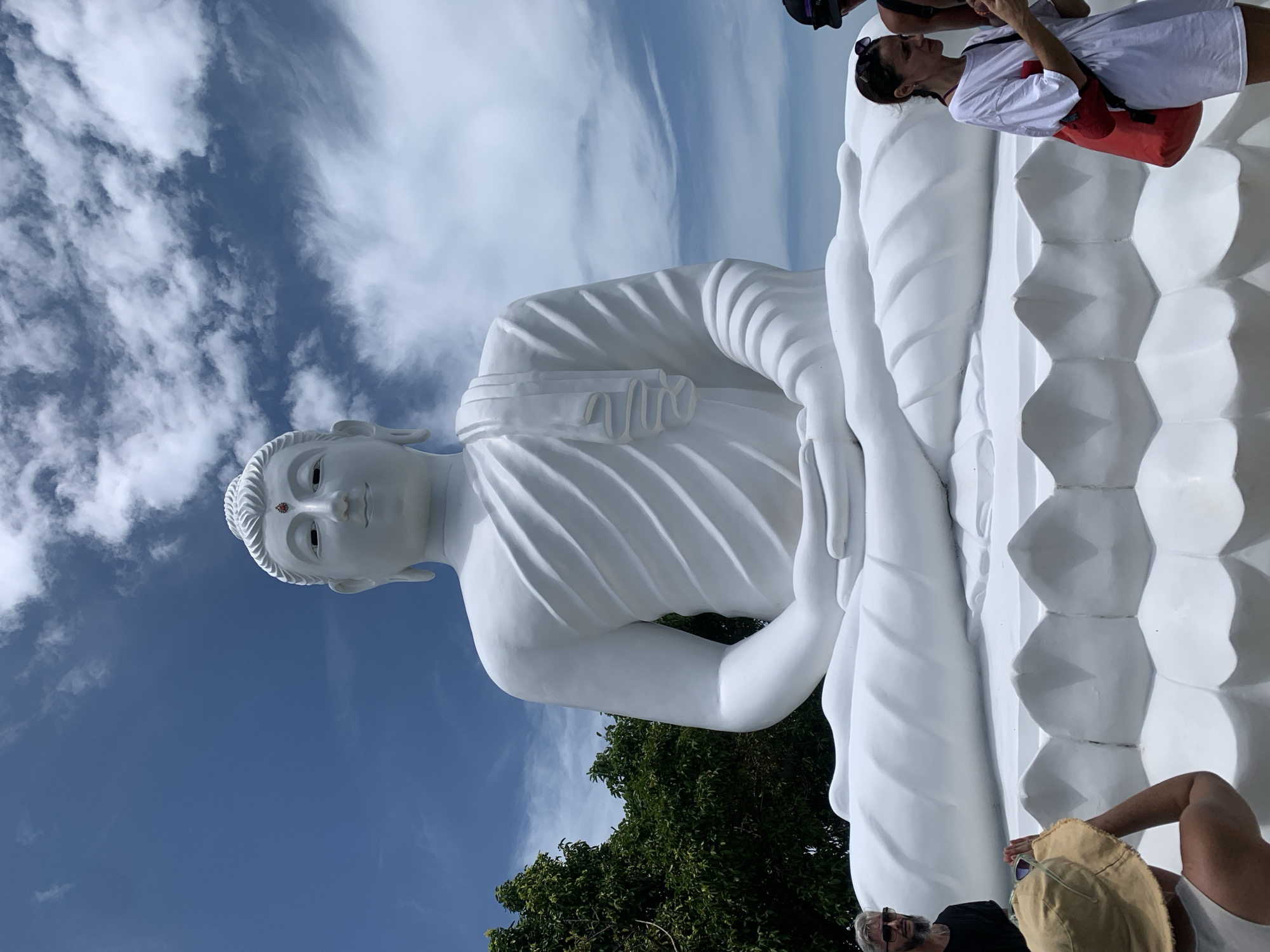 A large, white, seated Buddha statue dominates the frame against a bright blue sky with scattered clouds. The statue is detailed, with folds in its robe and a serene expression. In the foreground, several people are visible, appearing to be tourists. To the left, a man with a beard and sunglasses stands near the base of the statue. In the center, a person wearing a straw hat is partially visible from behind. To the right, a woman in a white t-shirt and a red bag stands looking at her phone. Another person in a black hat is partially visible to the far right. Lush green trees are visible behind the statue on the left side. The scene appears to be an outdoor tourist attraction, likely in Thailand given the cultural context of a large Buddha statue and tropical foliage.
