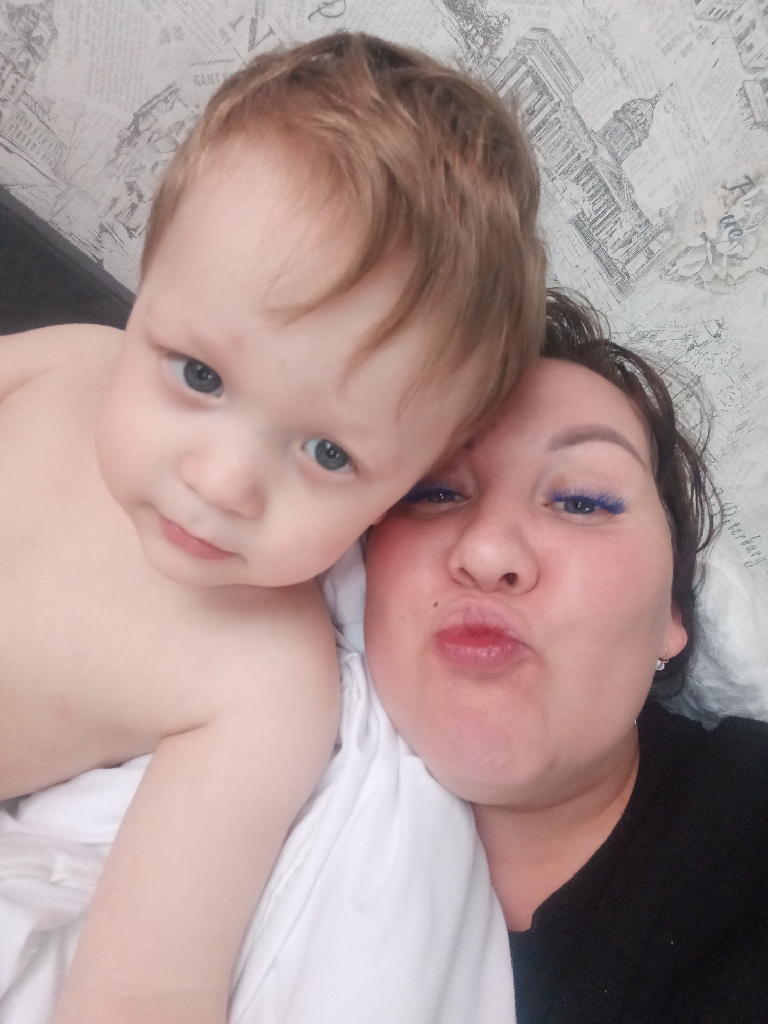 The image features a woman and a young child, likely a mother and her baby or toddler, captured in a close-up, high-angle selfie. The woman is on the right side of the frame, with dark hair and blue mascara on her eyelashes. She sports a small, clear earring and a mole on her right cheek, and wears a black shirt. Her lips are pursed in a playful "kiss face" expression.
To her left, partially covering her, is a fair-skinned child with light brown hair and prominent blue eyes, looking directly at the camera with a neutral or slightly curious expression. The child is shirtless, showing their bare chest and arm. Both individuals are lying down, resting on or against white bedding or fabric.
The background is a light-toned wallpaper adorned with black-and-white architectural sketches, depicting classical buildings with domes and columns, and interspersed with text. Visible words on the wallpaper include "GANT" and, along the right edge, "Peterburg" written vertically. These details, especially "Peterburg" (referring to St. Petersburg), align with the provided location context of Osa, Russia. The lighting is soft and even, indicative of an indoor setting.