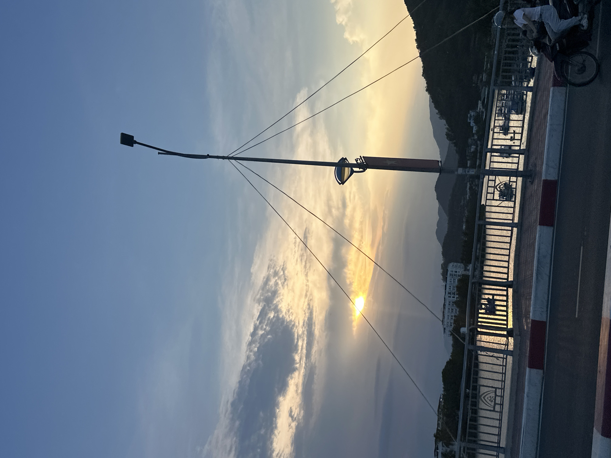 The image depicts a scene at sunset in Bac Nha Trang Ward, Vietnam. A long, thin lamppost with a black light fixture extends across the frame from left to right, anchored by several supporting wires. A metal and concrete bridge with red and white painted barriers spans a body of water, reflecting the setting sun's golden light. On the bridge, a person rides a motorbike. Distant mountains are visible beyond the water, along with some buildings. The sky above is a blend of light blue and golden hues, partially obscured by clouds.