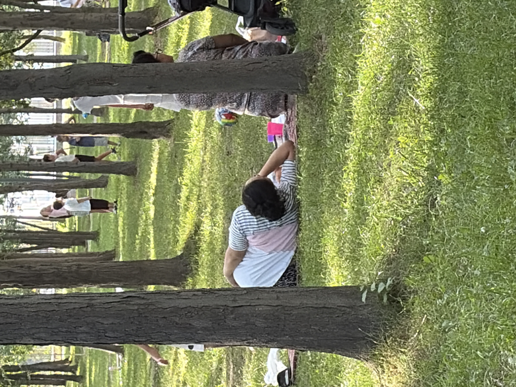 In an outdoor park setting, a person is lying on their side on a blanket, resting their head on their hand. The person is wearing a striped shirt and white pants, and their hair is pulled back. In the background, other people, including children, are visible walking and standing among the trees. A stroller is parked nearby. The ground is covered in grass, and sunlight filters through the trees, creating dappled shadows. The scene appears to be a relaxed day in a park, with individuals enjoying the outdoors.