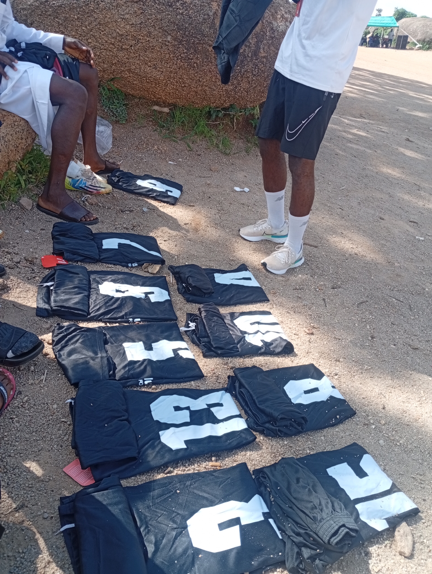 A group of dark t-shirts with white numbers and some dark shorts are spread out on the dusty ground near a large rock formation. Two individuals are partially visible, one sitting and wearing a white shirt and white shorts, and the other standing and wearing a white shirt and black shorts. The scene appears to be outdoors, with sparse vegetation and sand. This image was taken in Jos, Nigeria.