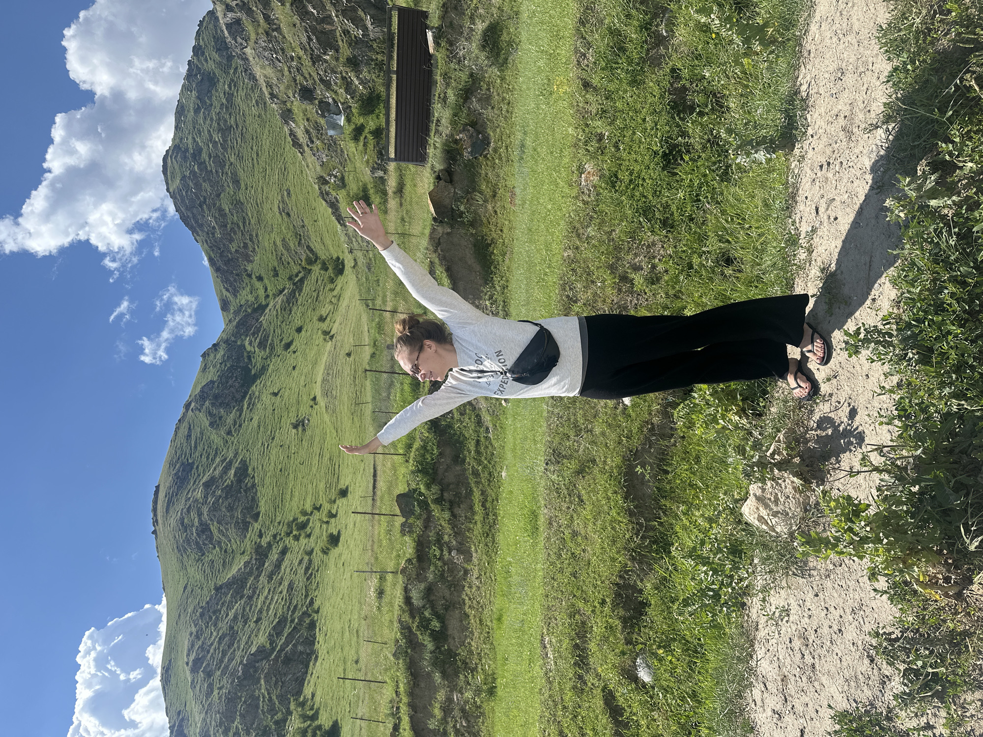 A woman is standing on a dirt path in a grassy, mountainous landscape. She is wearing a light-colored long-sleeved shirt, black pants, and flip-flops. A black fanny pack is worn across her chest. Her arms are outstretched, and she appears to be joyful or exhilarated. The background features a steep, green mountain under a bright blue sky with scattered white clouds. A simple metal structure, possibly a bench, is visible on the mountainside. The scene suggests a sunny day in a rural, possibly remote, location. The image is oriented vertically. There is visible text on the woman's shirt, but it is largely illegible due to resolution and angle.